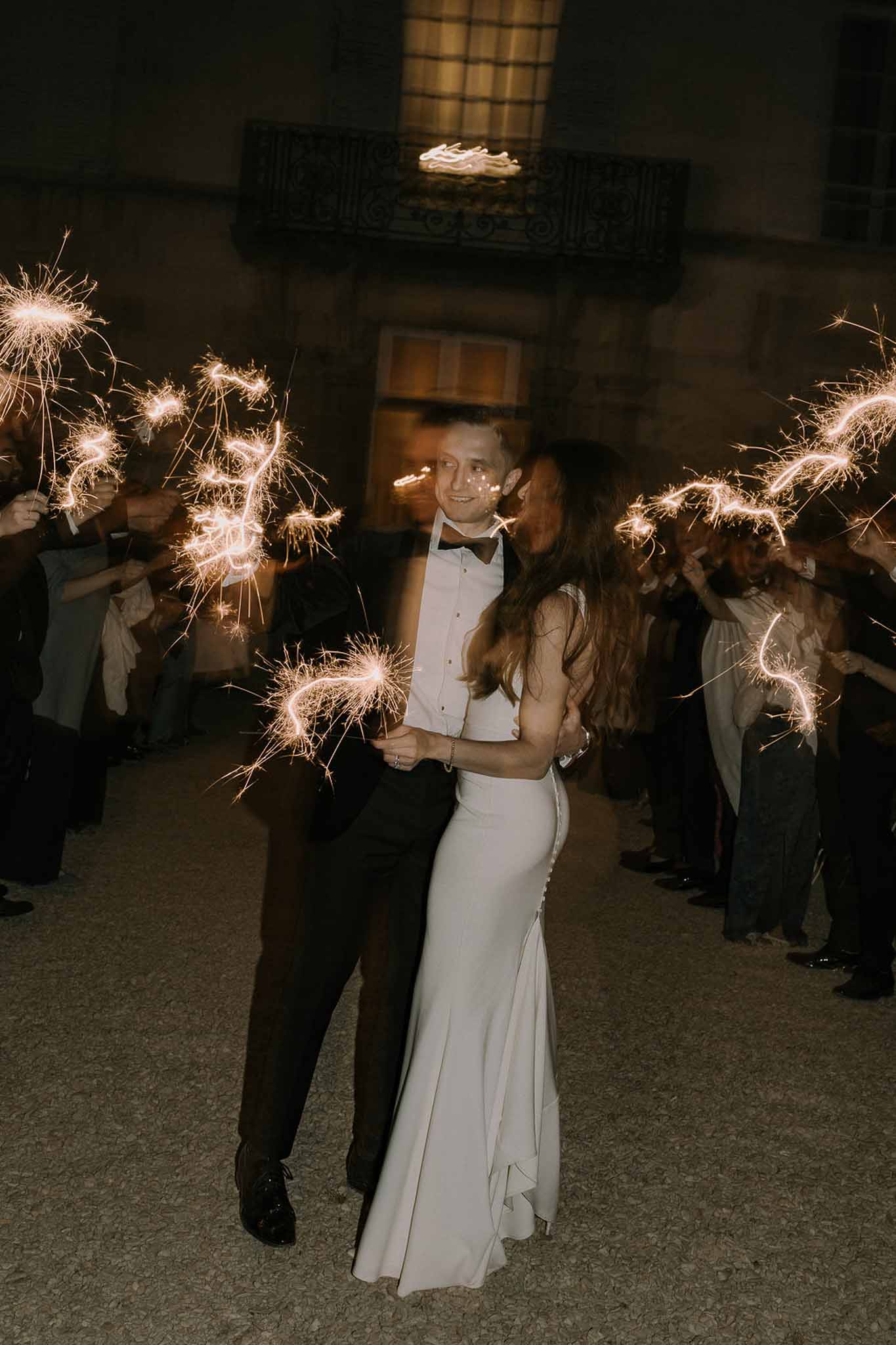 Bride and groom walk through sparkler send-off tunnel held by guests on gravel path at night