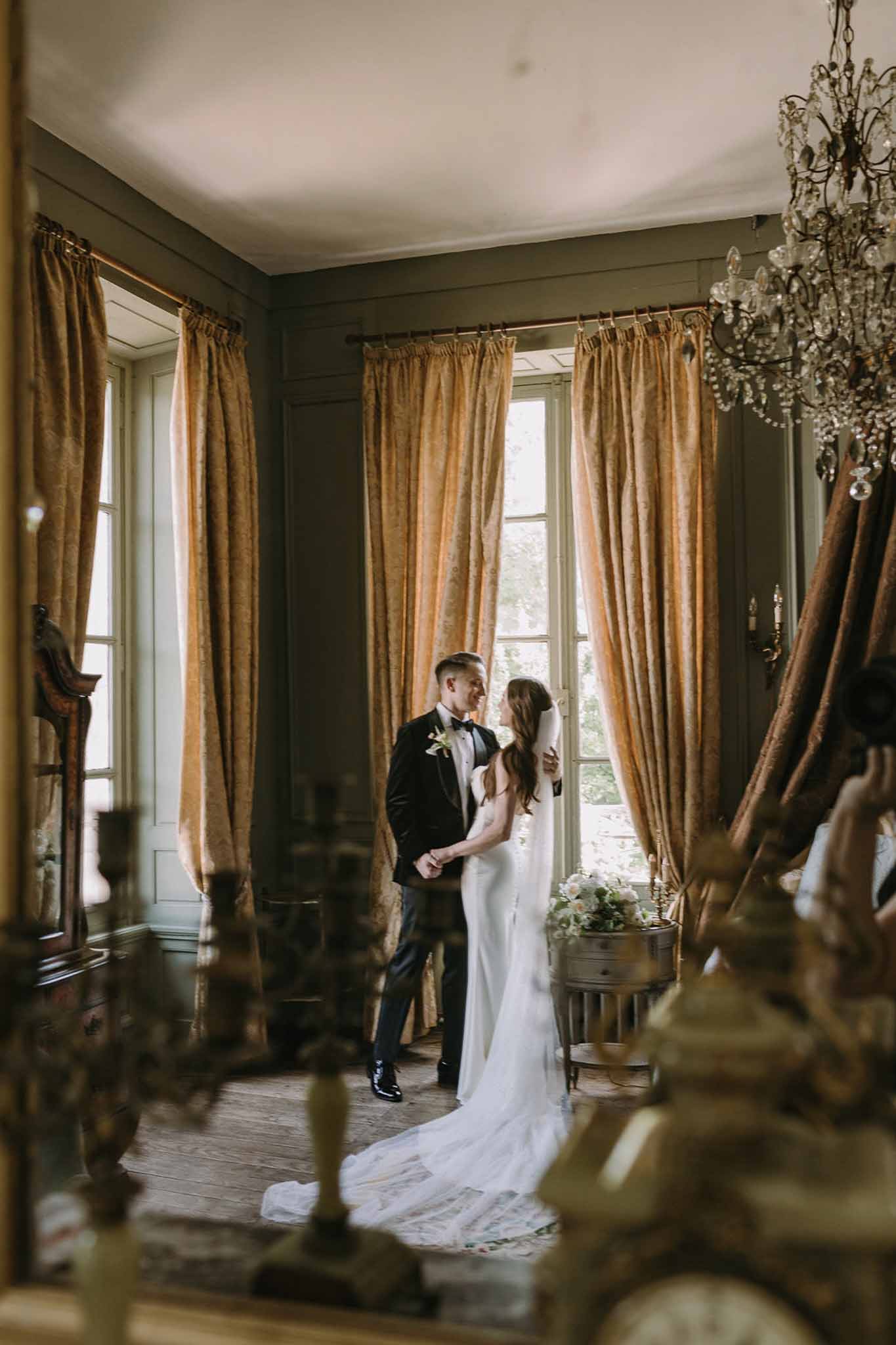 Bride and groom holding hands in chateau room with olive-green panels, gold curtains, and chandelier