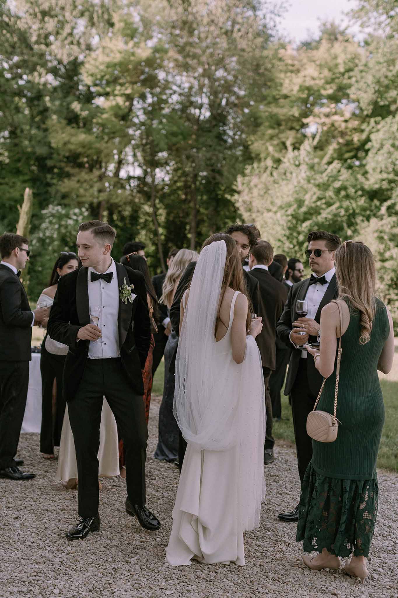 Bride and groom mingling with black-tie guests during outdoor cocktail hour on gravel terrace under tall trees