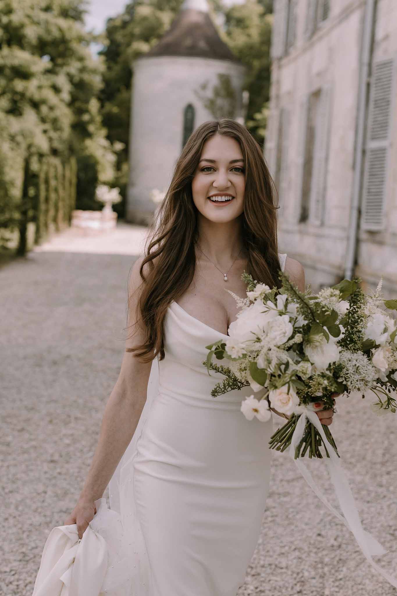 Smiling bride in cowl-neck slip gown holding white rose and anemone bouquet on chateau driveway