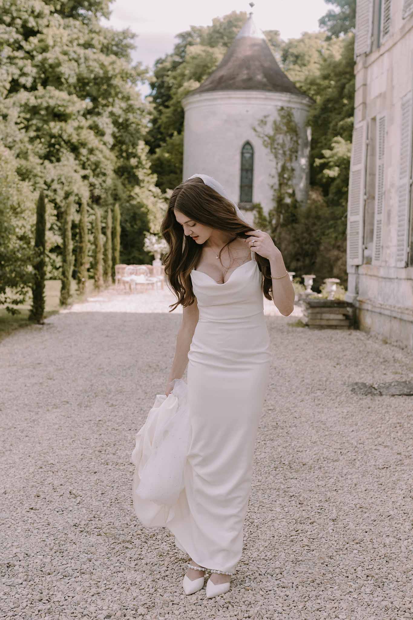 Bride in ivory satin column gown with detachable train and short veil beside chateau round tower