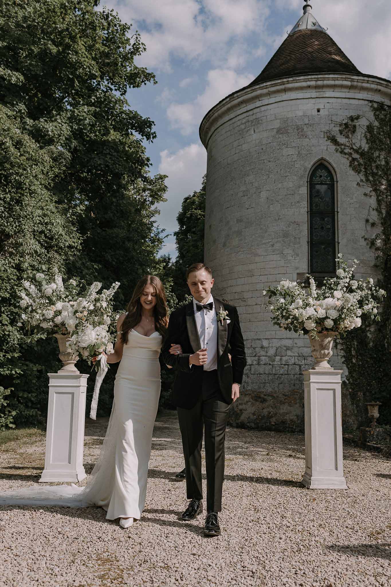 Bride and groom walking arm-in-arm past stone chateau tower flanked by white floral pedestal urns