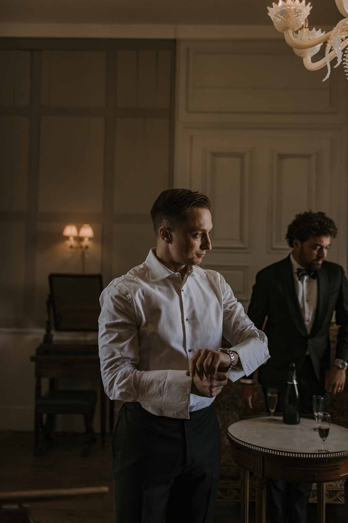 Groom adjusting watch cufflink with groomsman and champagne in boiserie-panelled room