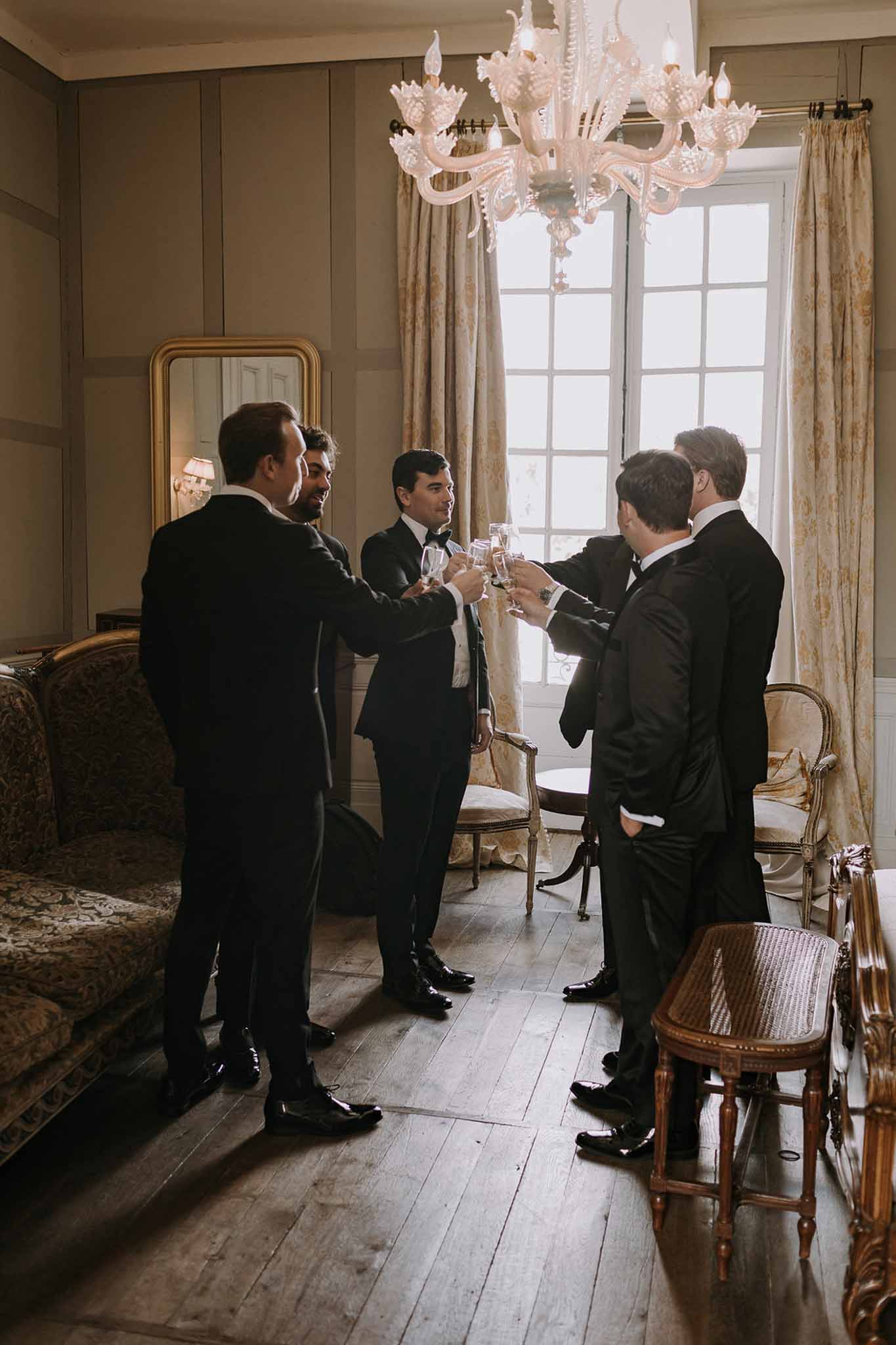 Five men in black tuxedos toast champagne in chateau salon with herringbone floors and Murano chandelier
