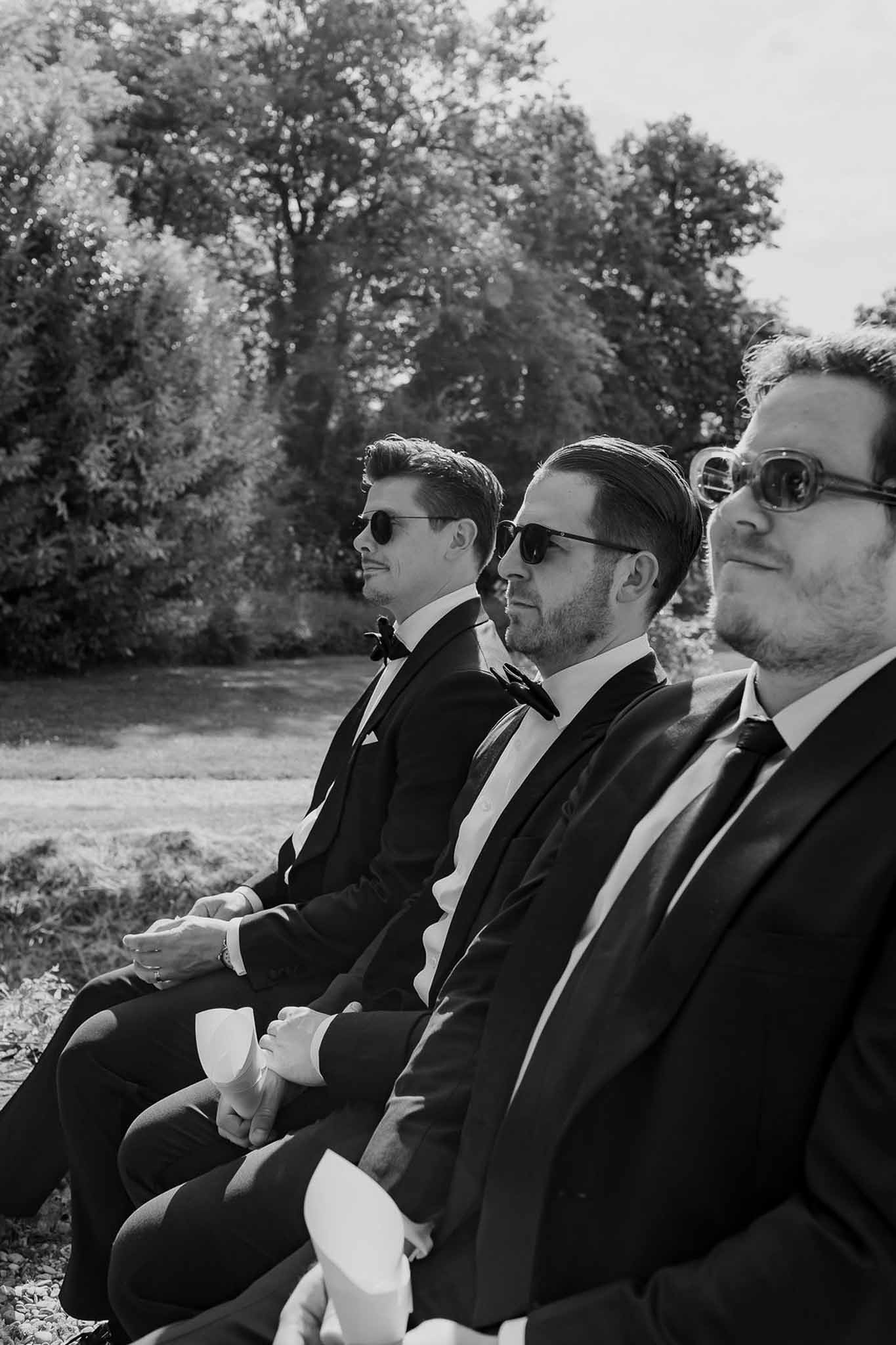 Black and white three groomsmen in dark suits and sunglasses seated watching outdoor garden ceremony