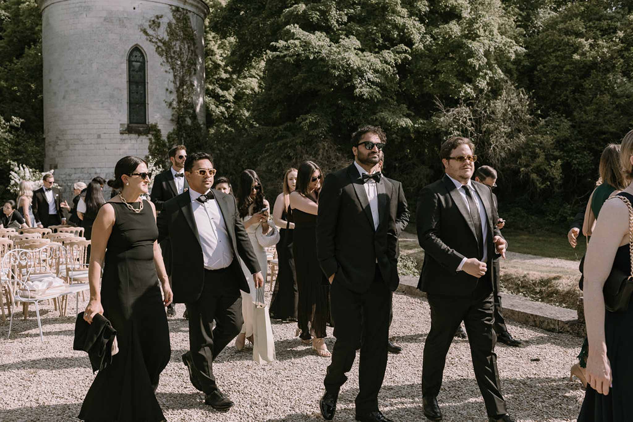 Guests in all-black formal dress code dispersing past white stone tower after outdoor ceremony