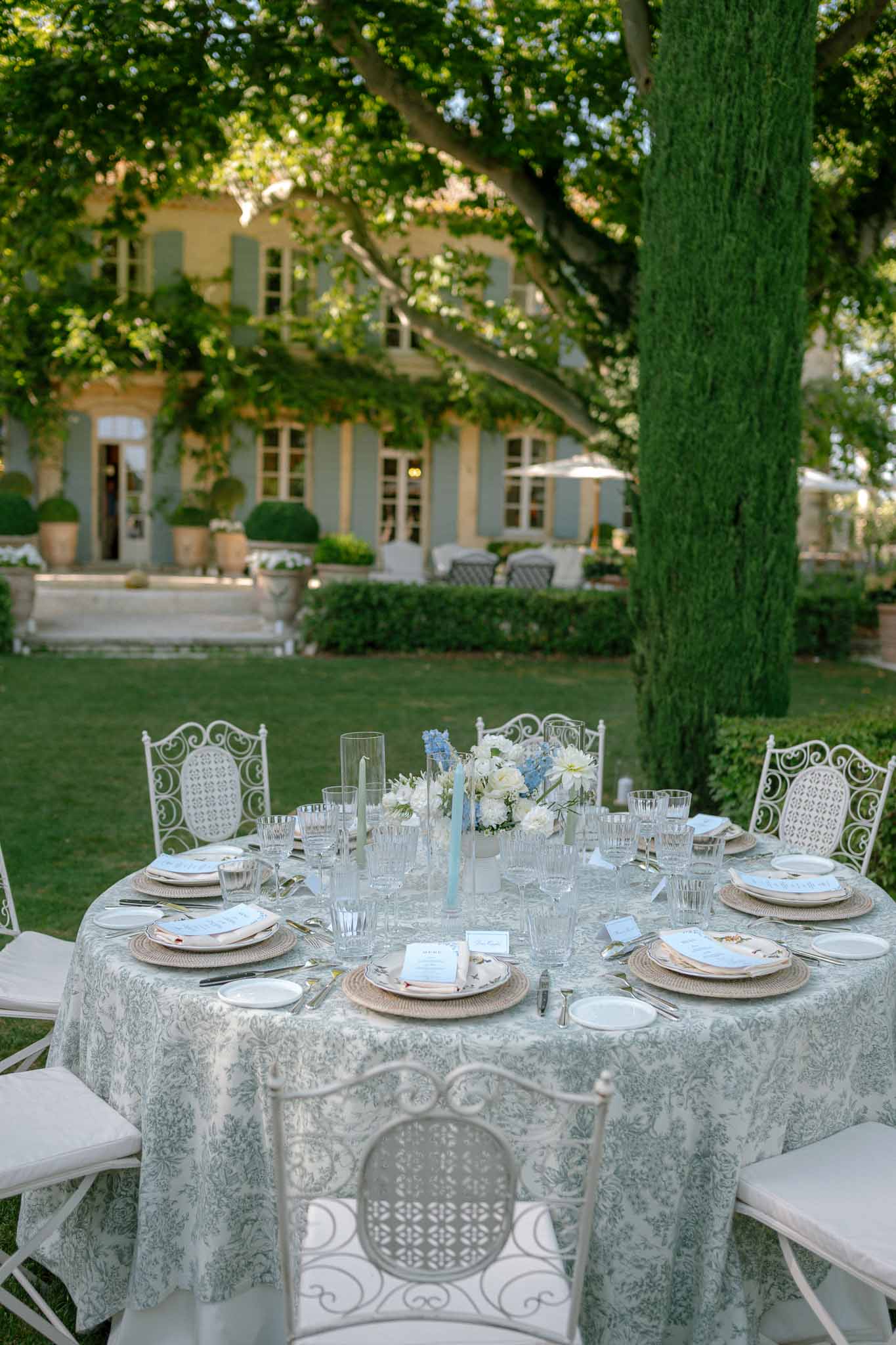 Round toile de Jouy table with blue hydrangea and white dahlia centerpiece before Provencal mas with sage shutters