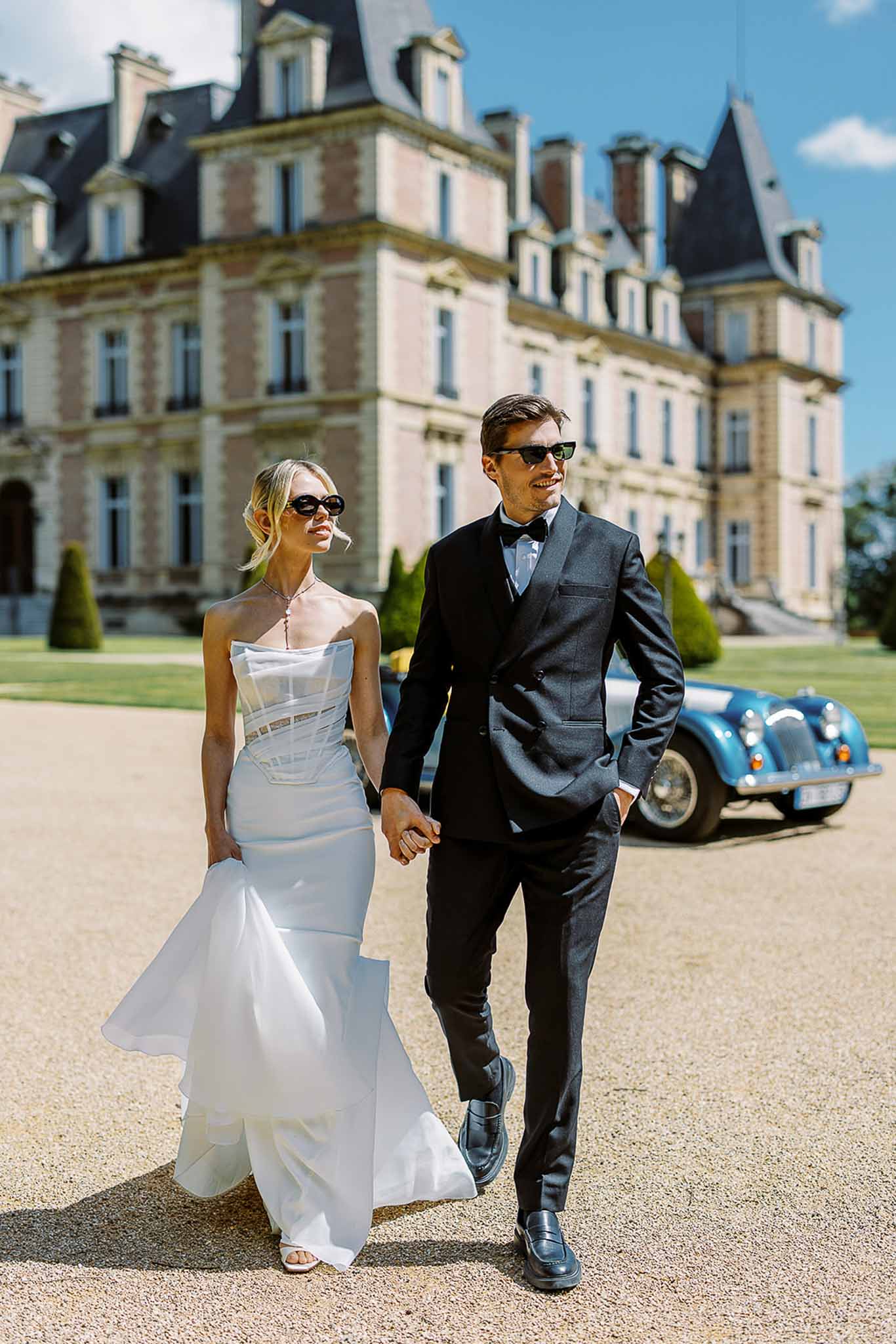Bride in strapless mermaid gown and groom in charcoal suit walking from vintage blue roadster at chateau