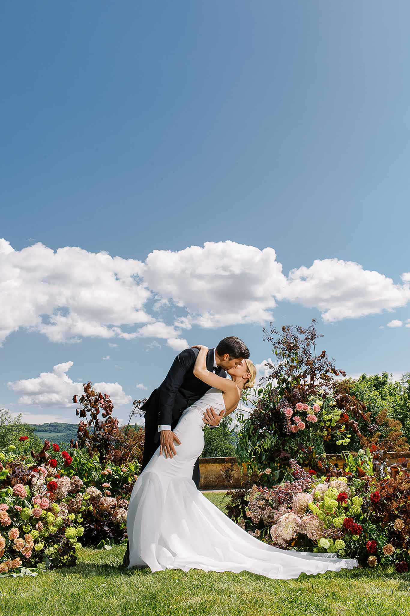 Groom dipping bride for kiss on lawn with burgundy dahlia and blush hydrangea garden border behind