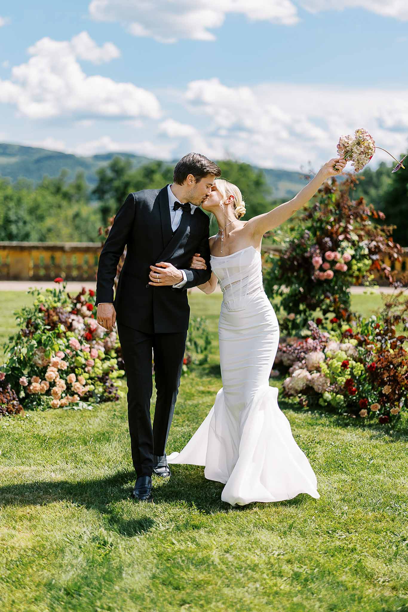 Bride and groom kissing on lawn with bride raising pink bouquet beside ground-level floral installations