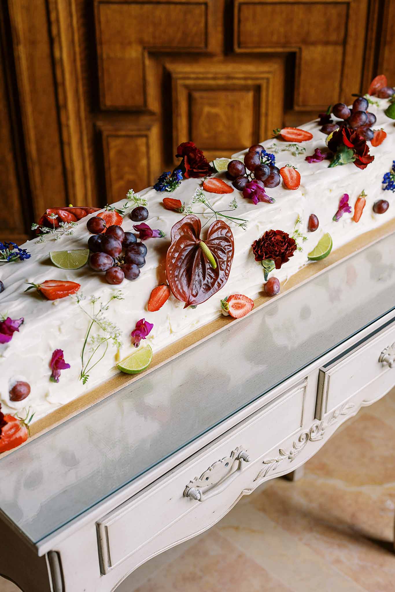 Elongated white cake decorated with grapes, strawberries, burgundy anthurium, and sweet peas on console