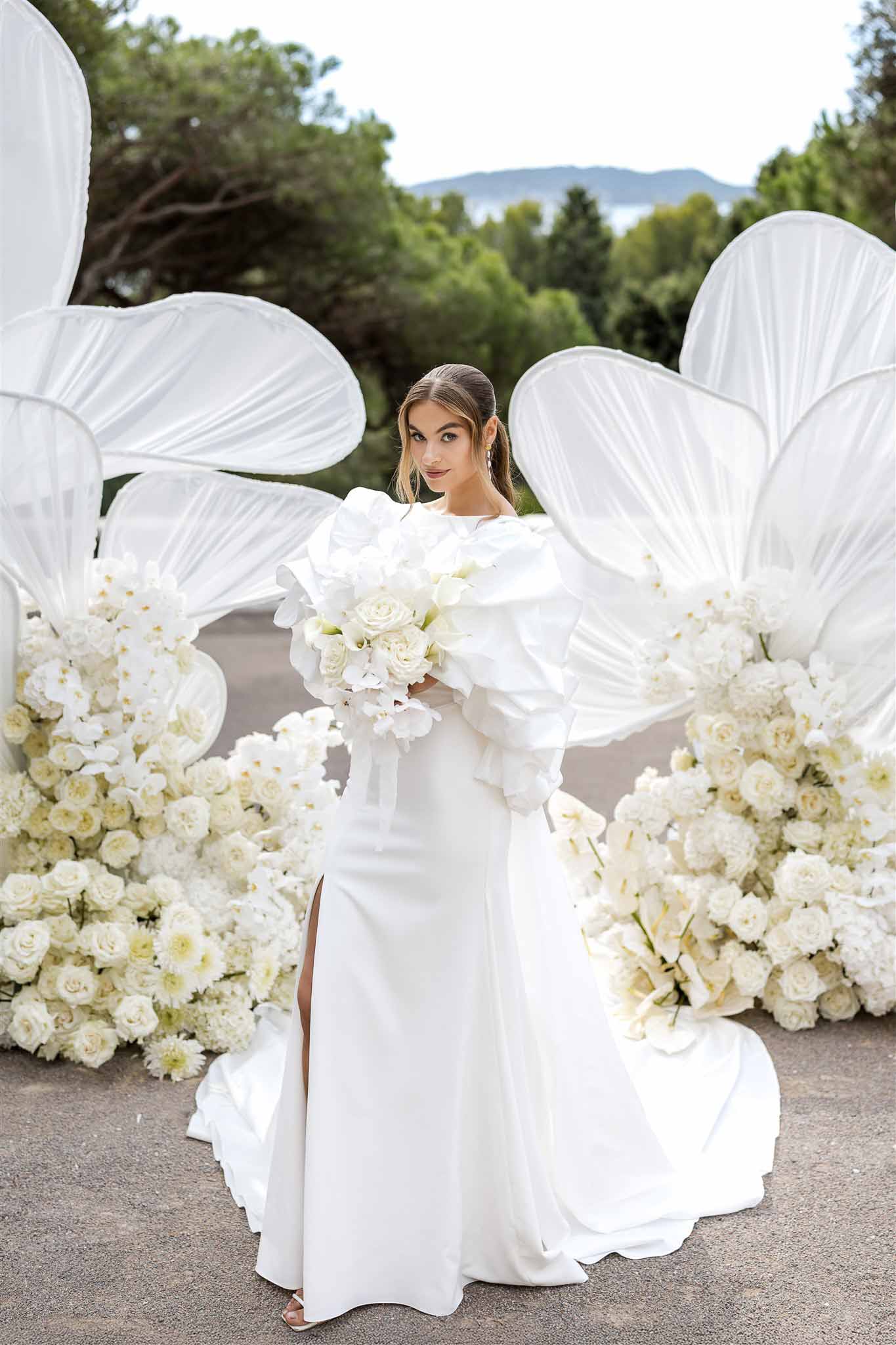 Bride in fitted gown with ruffle sleeves holding white orchid bouquet before sculptural petal installation