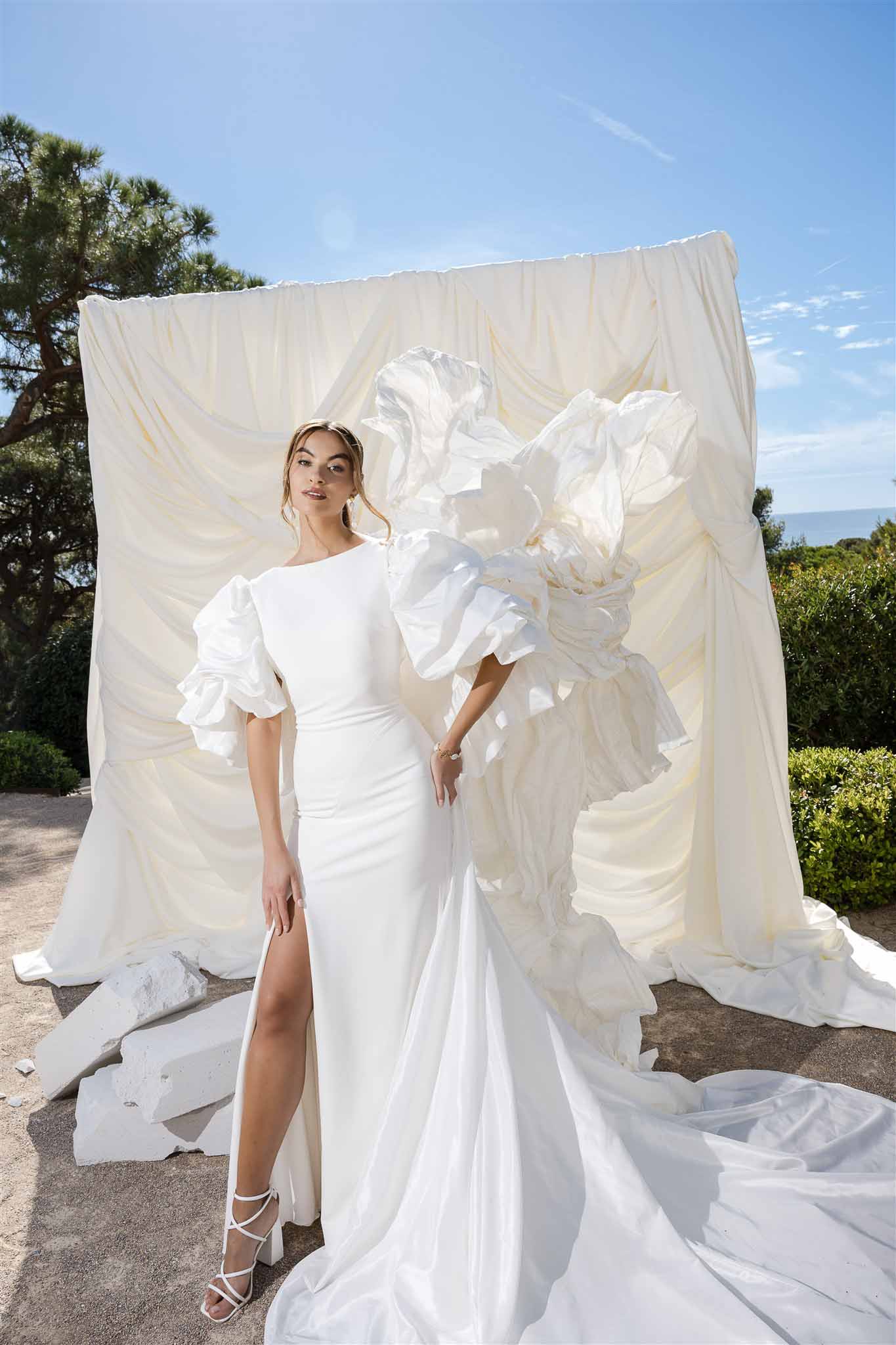 Bride in white crepe gown with dramatic puff sleeves and thigh slit before draped ivory backdrop by the sea