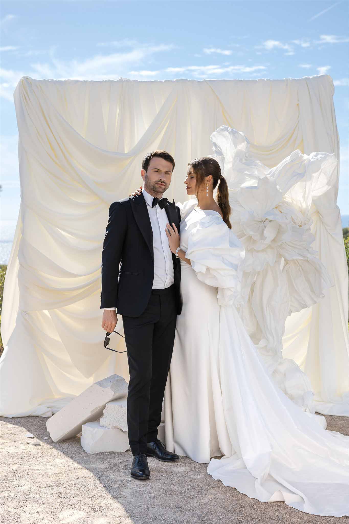 Couple before draped ivory backdrop with sculptural white florals, bride in puff-sleeve gown, coastal view behind