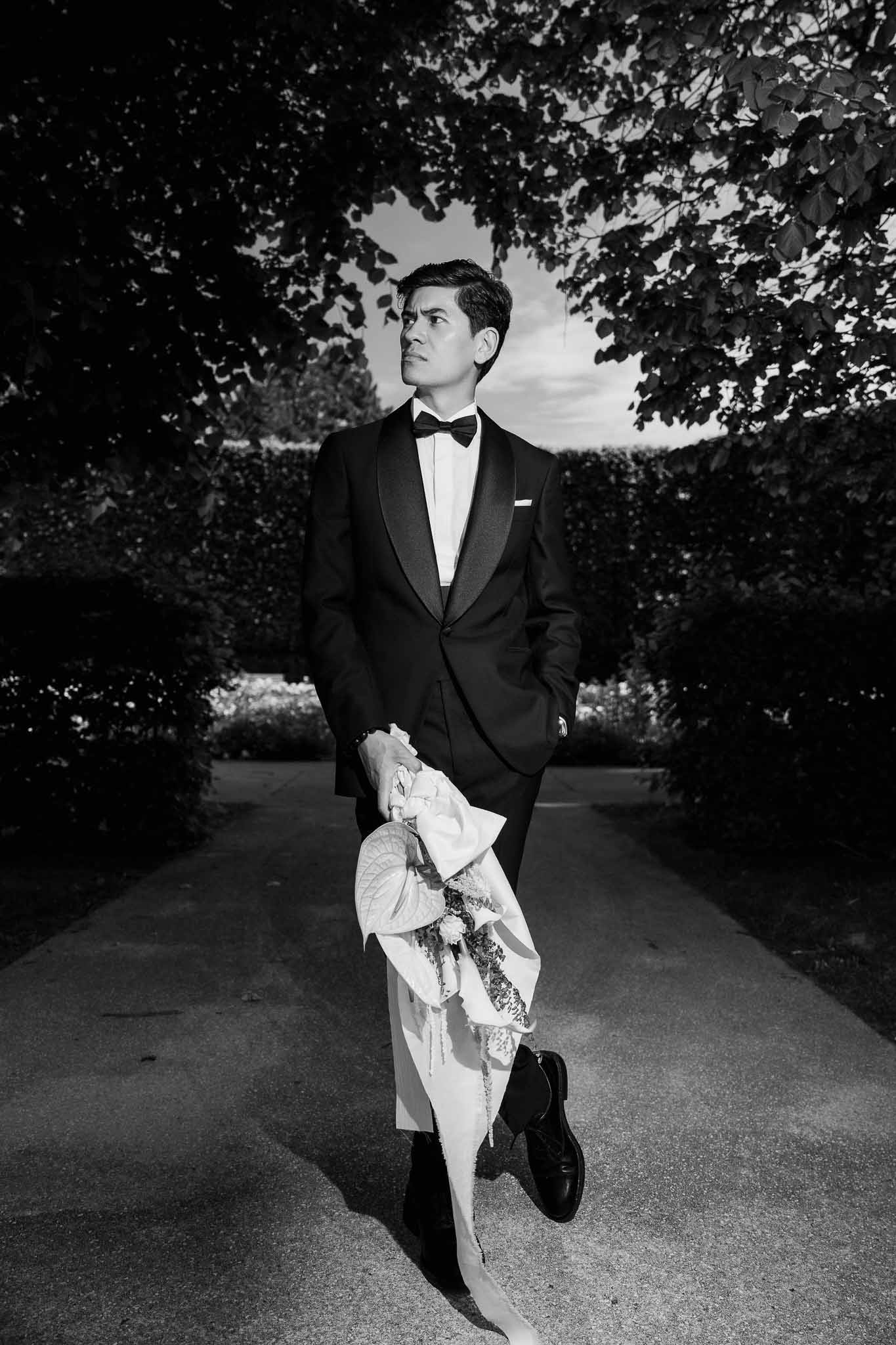 Black and white full-length portrait of groom in tuxedo holding bouquet on hedge-lined garden path