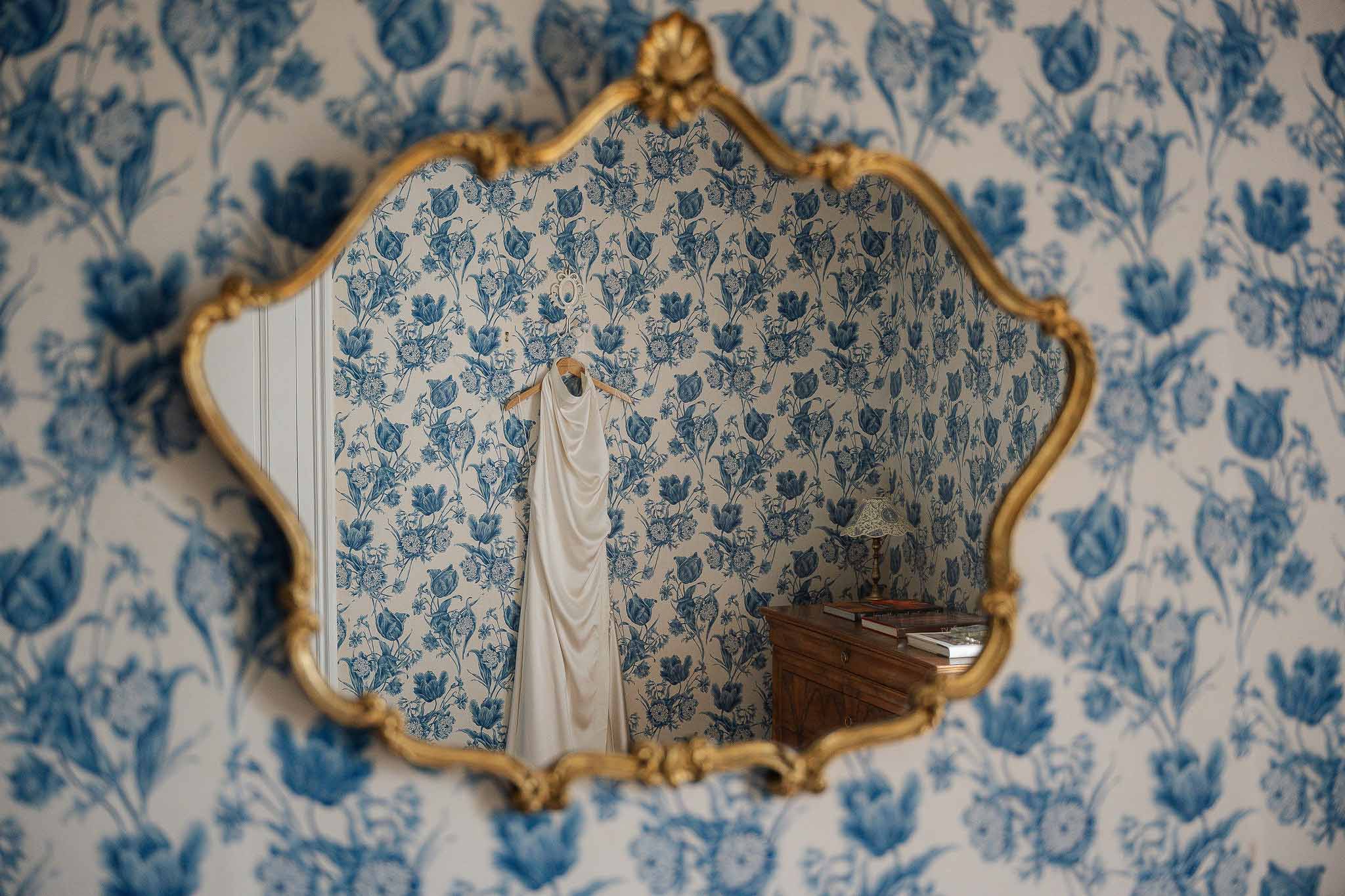 Ivory satin wedding dress reflected in gold-framed mirror on blue and white toile wallpaper in French interior