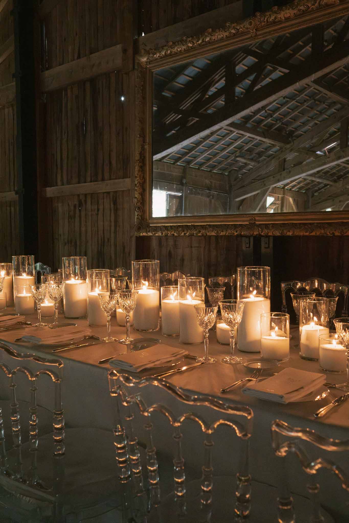 Rustic barn reception table with pillar candles, ghost chairs, and gilt mirror against timber walls