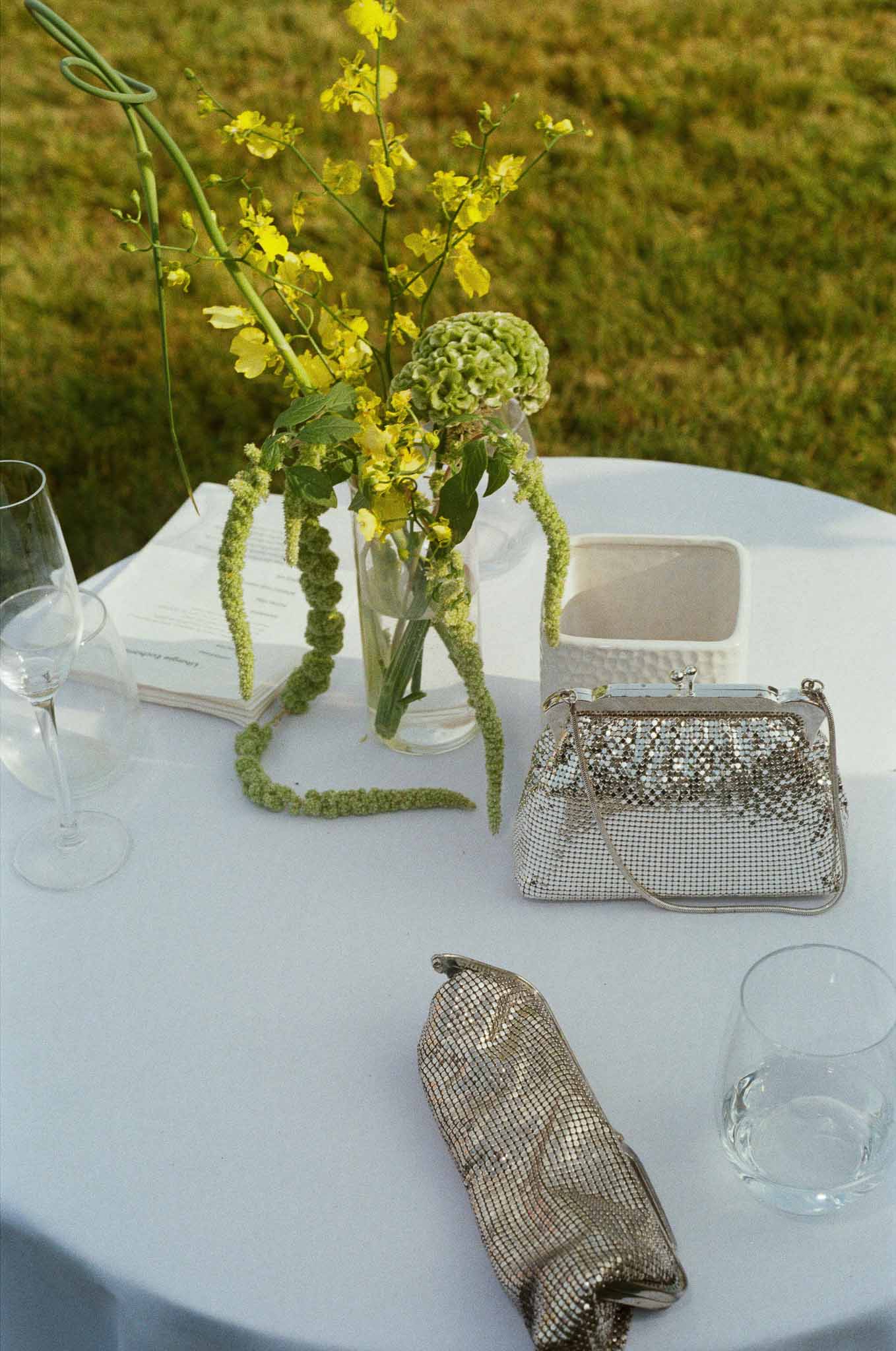 Cocktail table with yellow orchid and green hydrangea centerpiece, silver clutch bags, and wine glasses on white linen