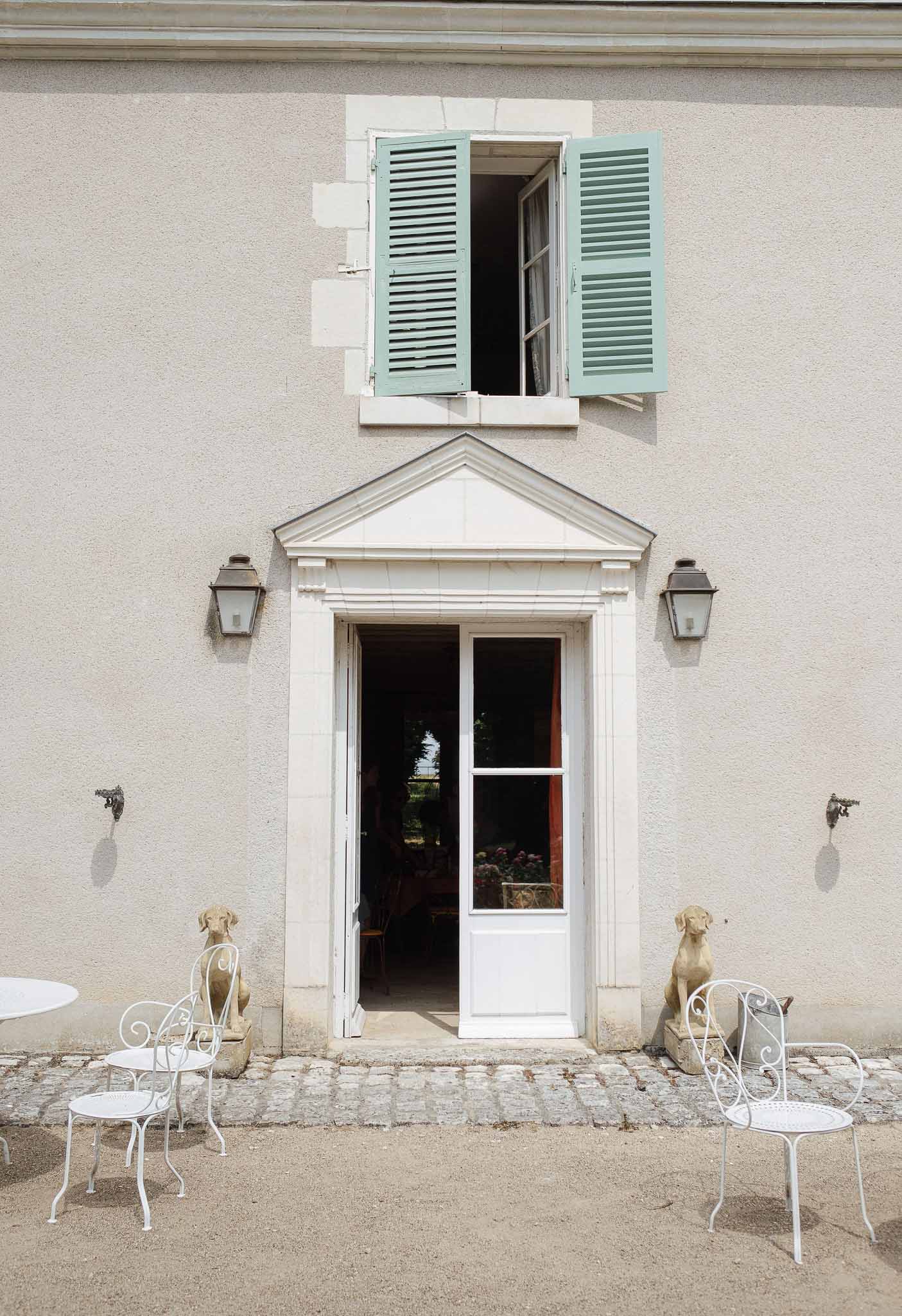 White-rendered French country house entrance with green shutters, stone dog statues, and bistro furniture