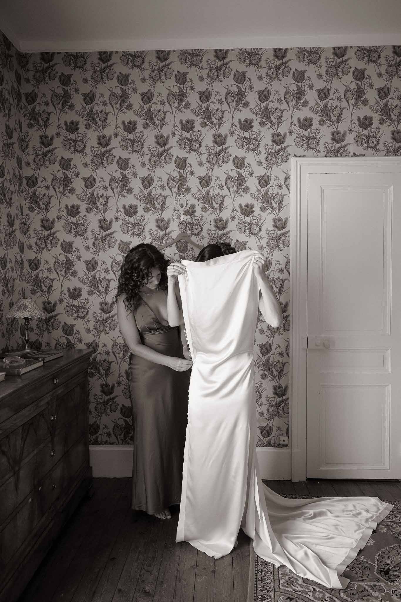 Black-and-white shot of bridesmaid helping bride into her wedding dress in a chateau bedroom with toile wallpaper