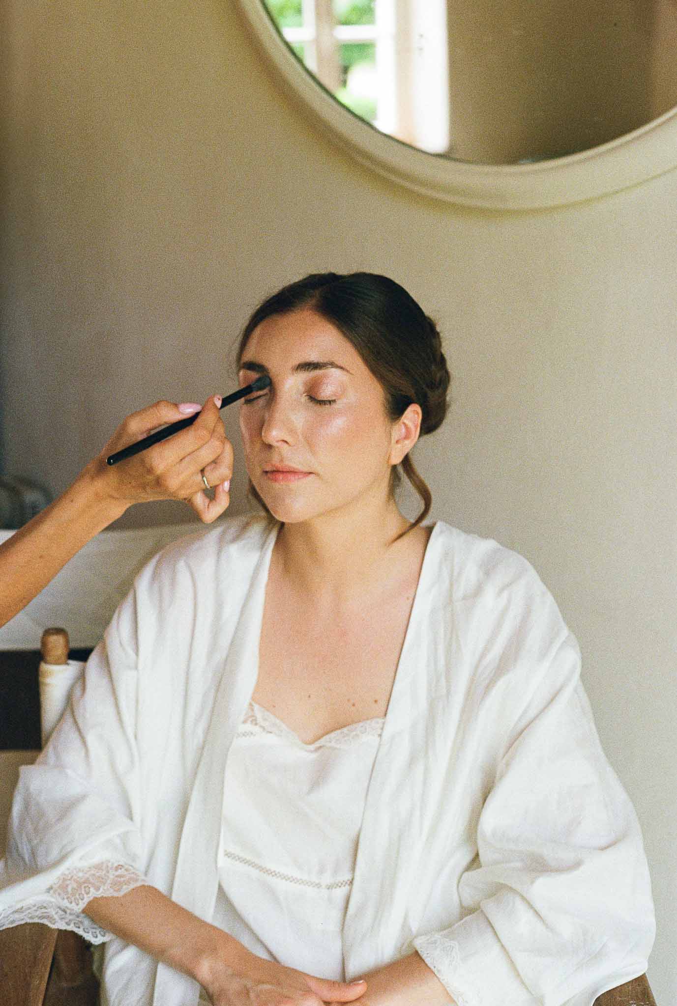 Bride in white robe having eyeshadow applied with brush in warm-toned room with gold-framed mirror