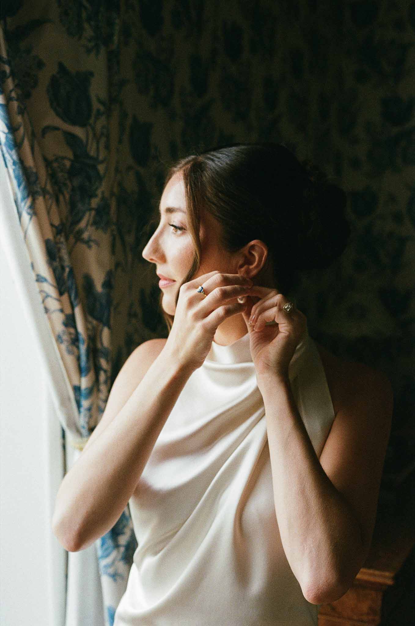 Bride fastening earring in ivory halter gown beside toile de Jouy curtains in natural window light