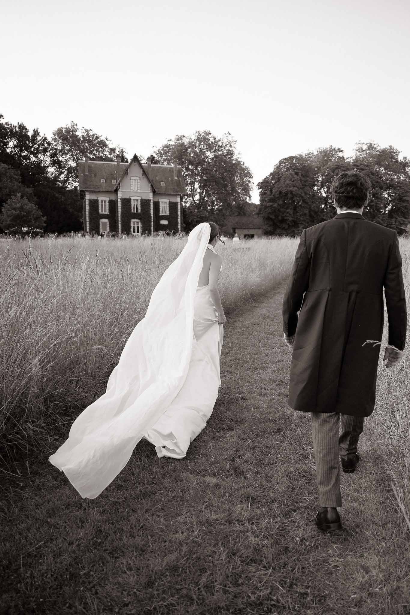 Black and white rear view of couple walking through meadow toward manor with billowing veil