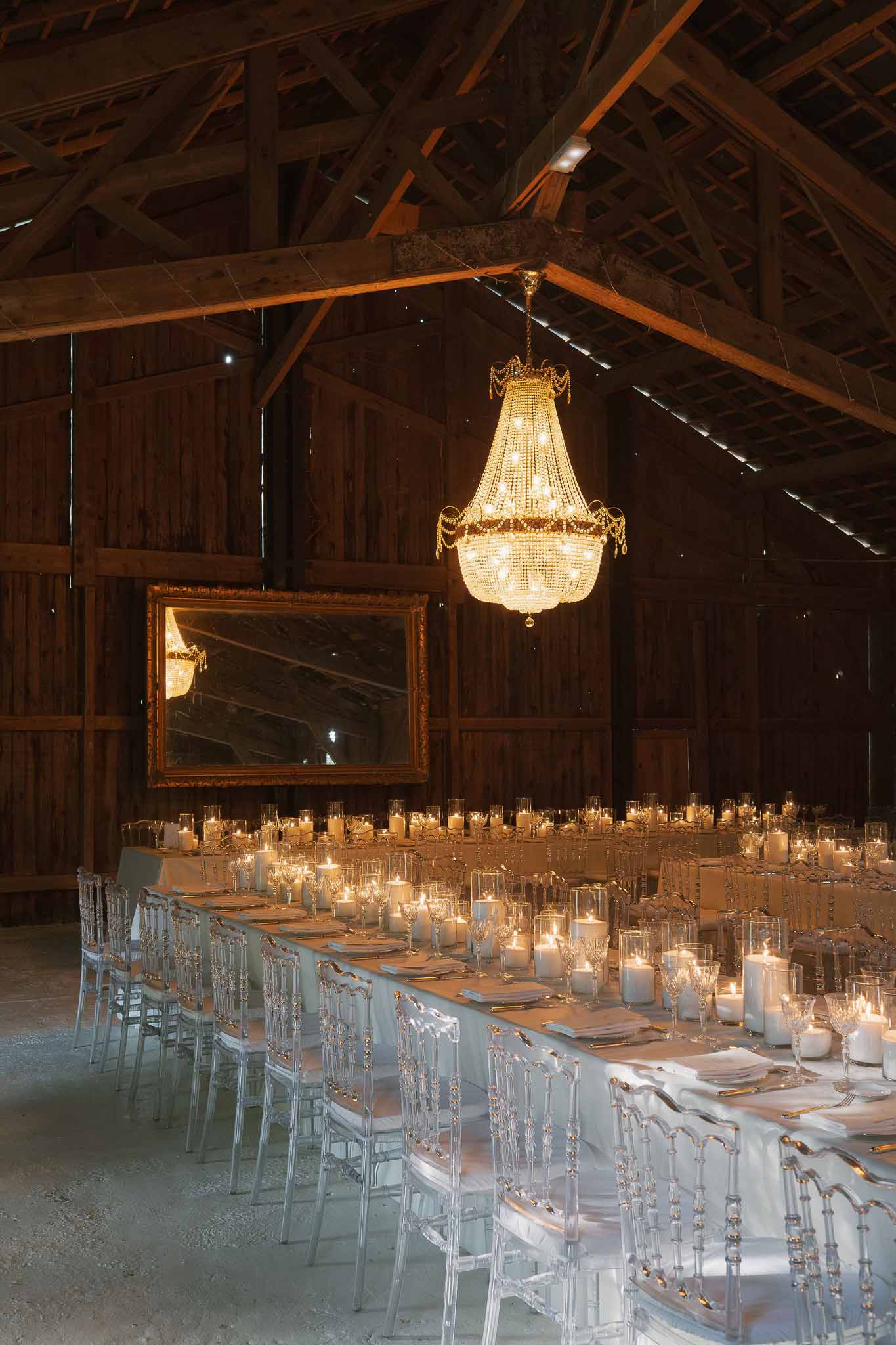 Rustic barn reception with gold crystal chandelier, ghost chairs, white pillar candles, and gold-framed mirror at back wall