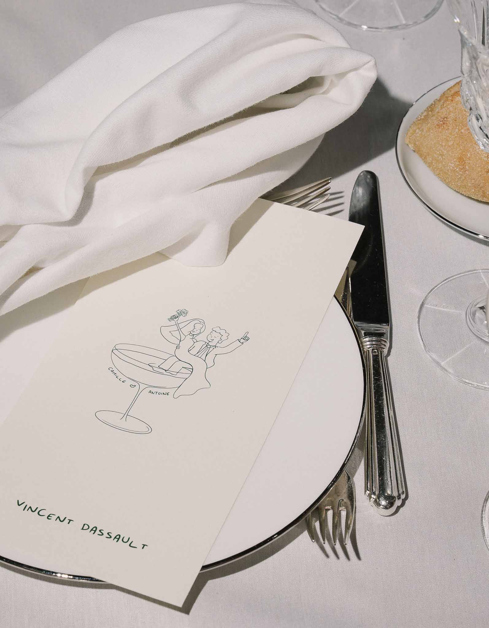 Silver-rimmed charger with illustrated champagne-coupe place card and crystal glasses on white linen