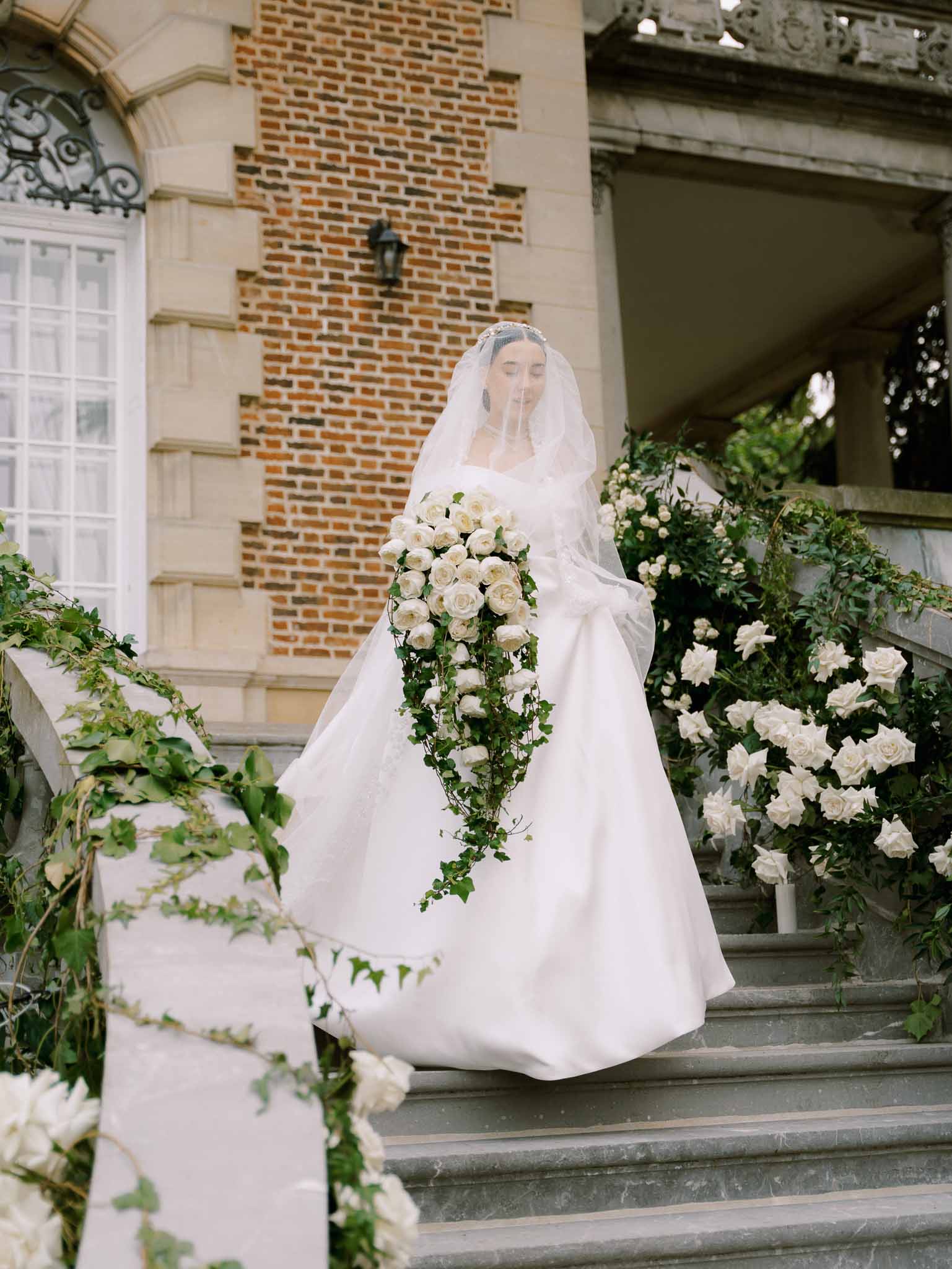 Bride in ivory ballgown with cathedral veil and cascading ivy bouquet on rose-draped chateau staircase
