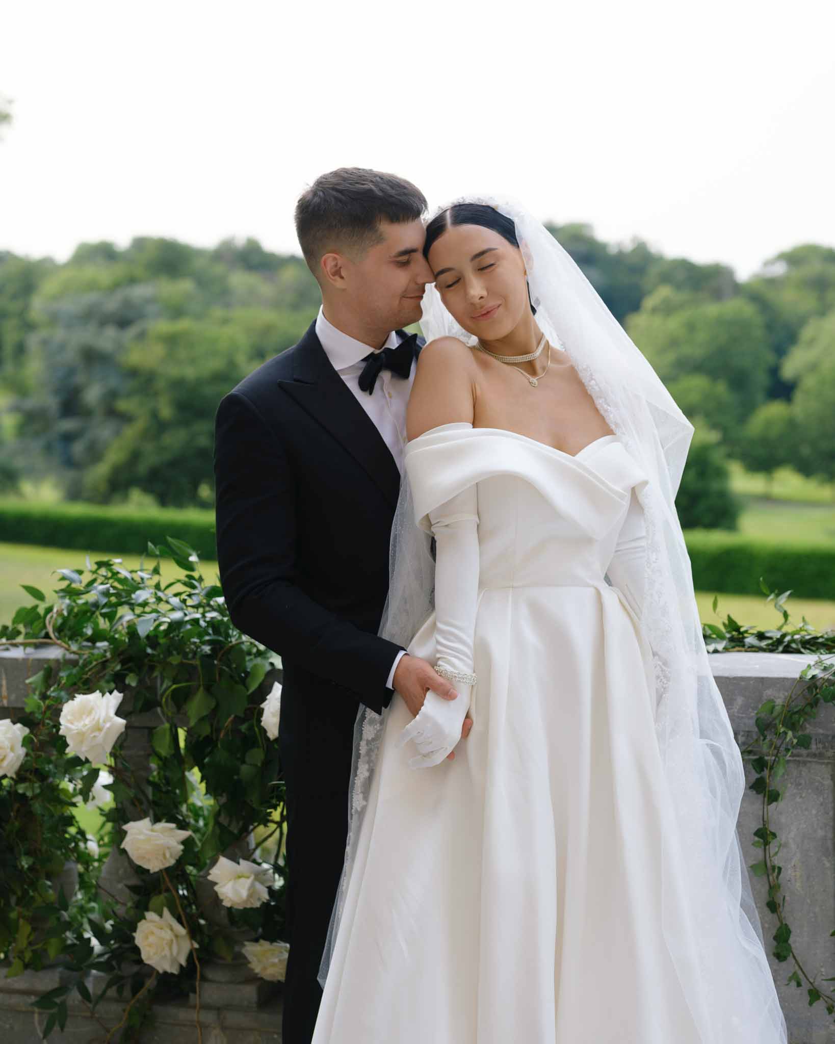 Bride and groom foreheads touching on stone terrace, bride in ballgown with lace gloves and pearl necklaces