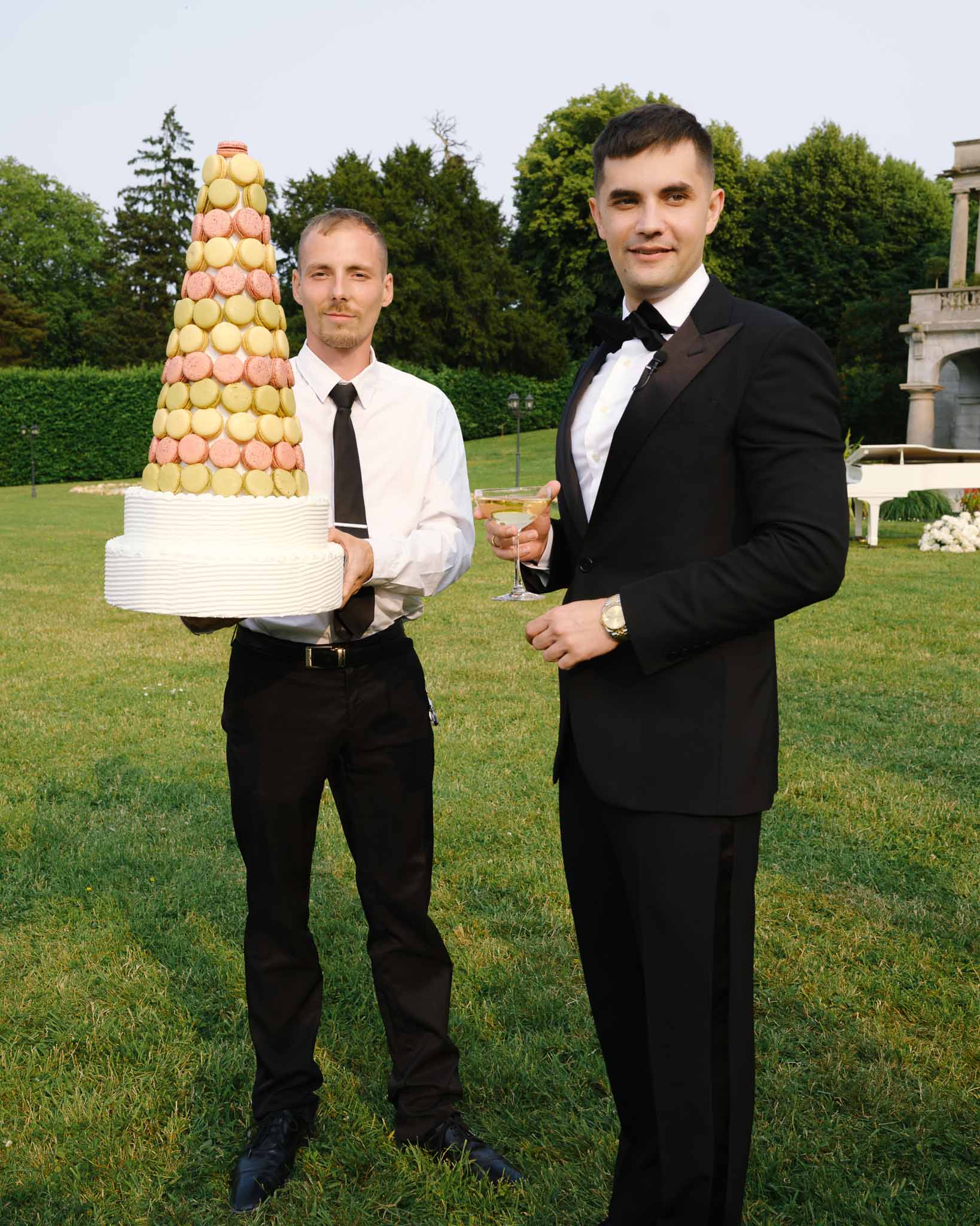 Staff member holding two-tiered white wedding cake with macaron tower beside groom on chateau lawn
