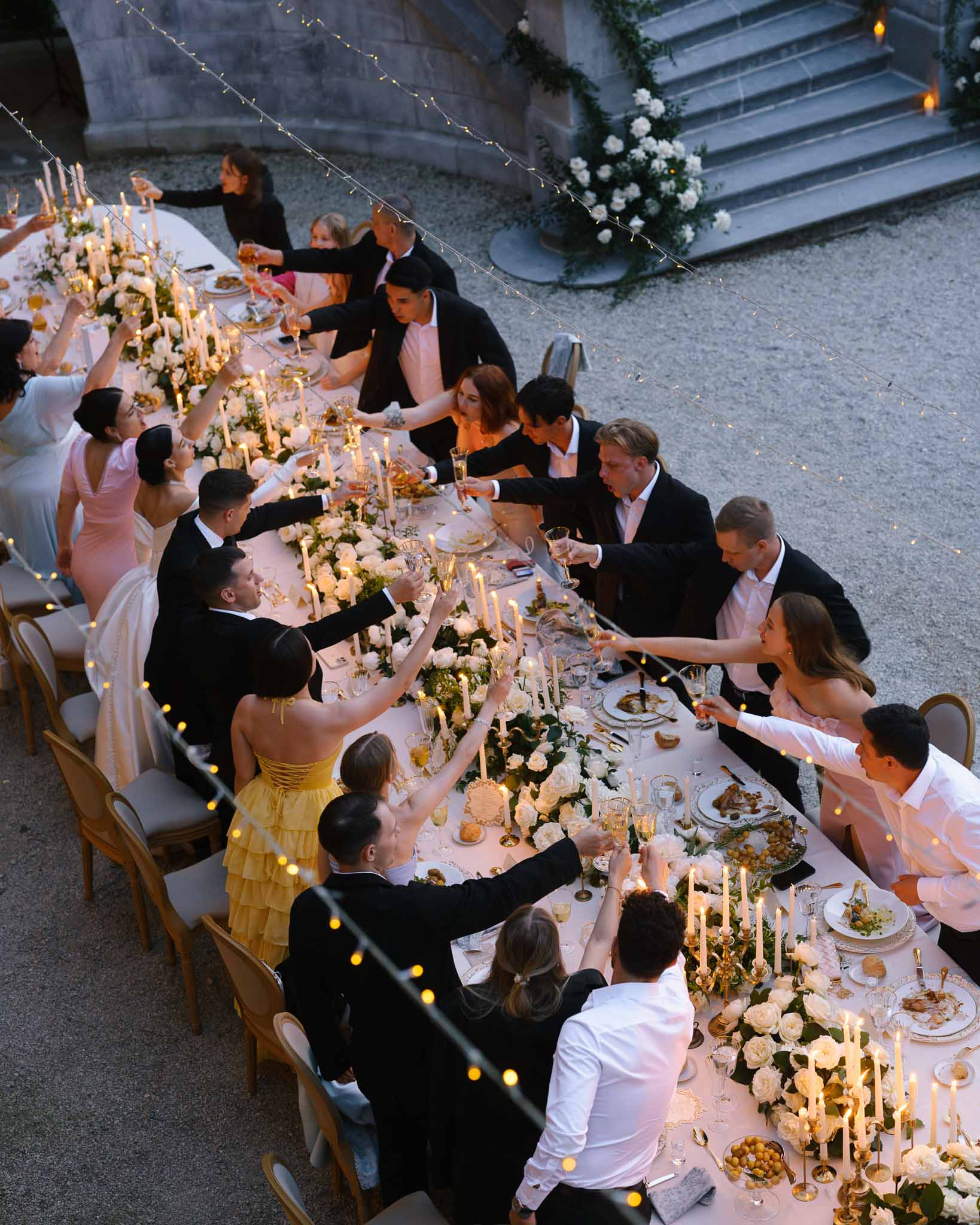Aerial view of guests raising flutes over ivory rose runner with gold candelabras at dusk on gravel
