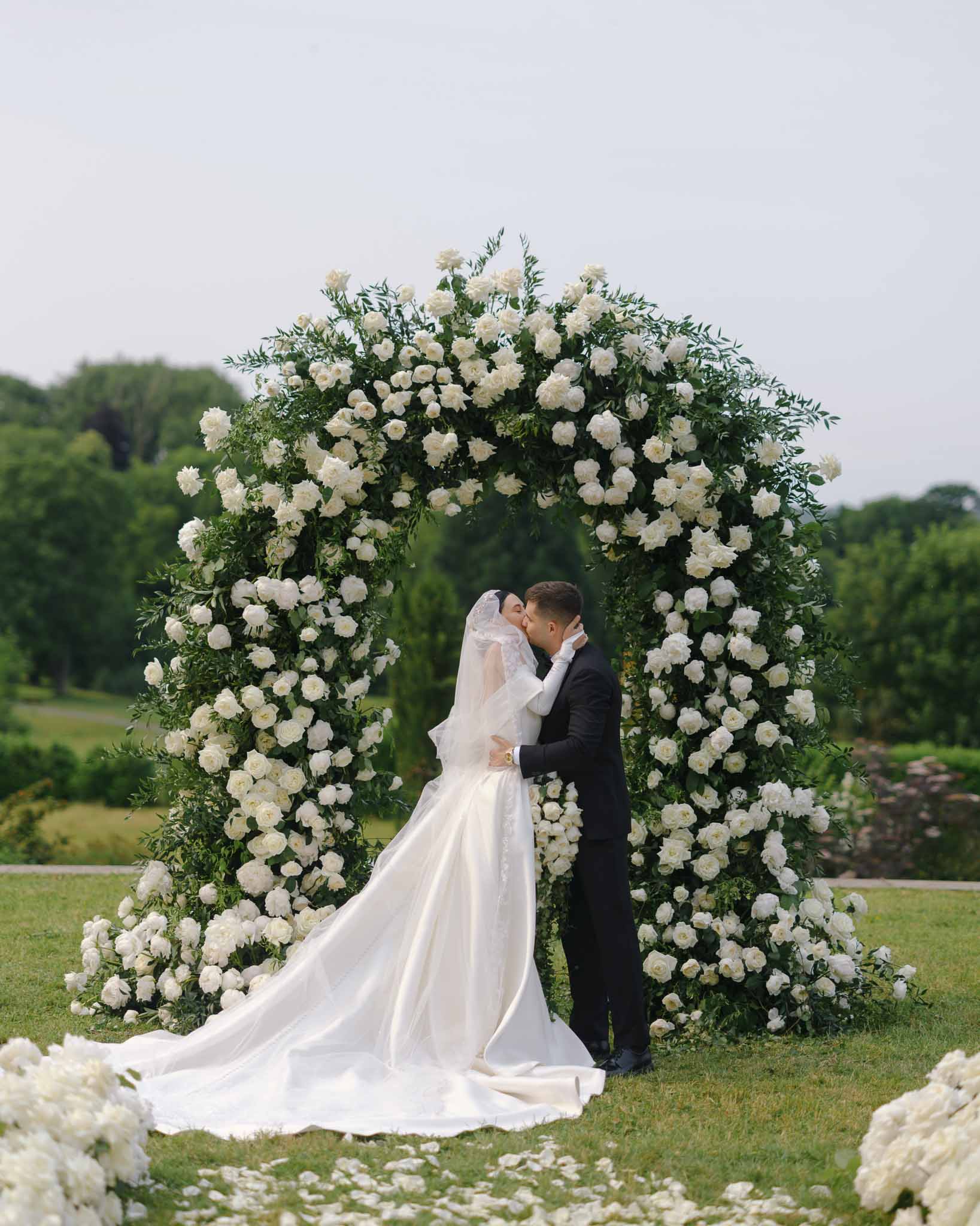 First kiss under circular white peony and rose arch with scattered petals on garden lawn