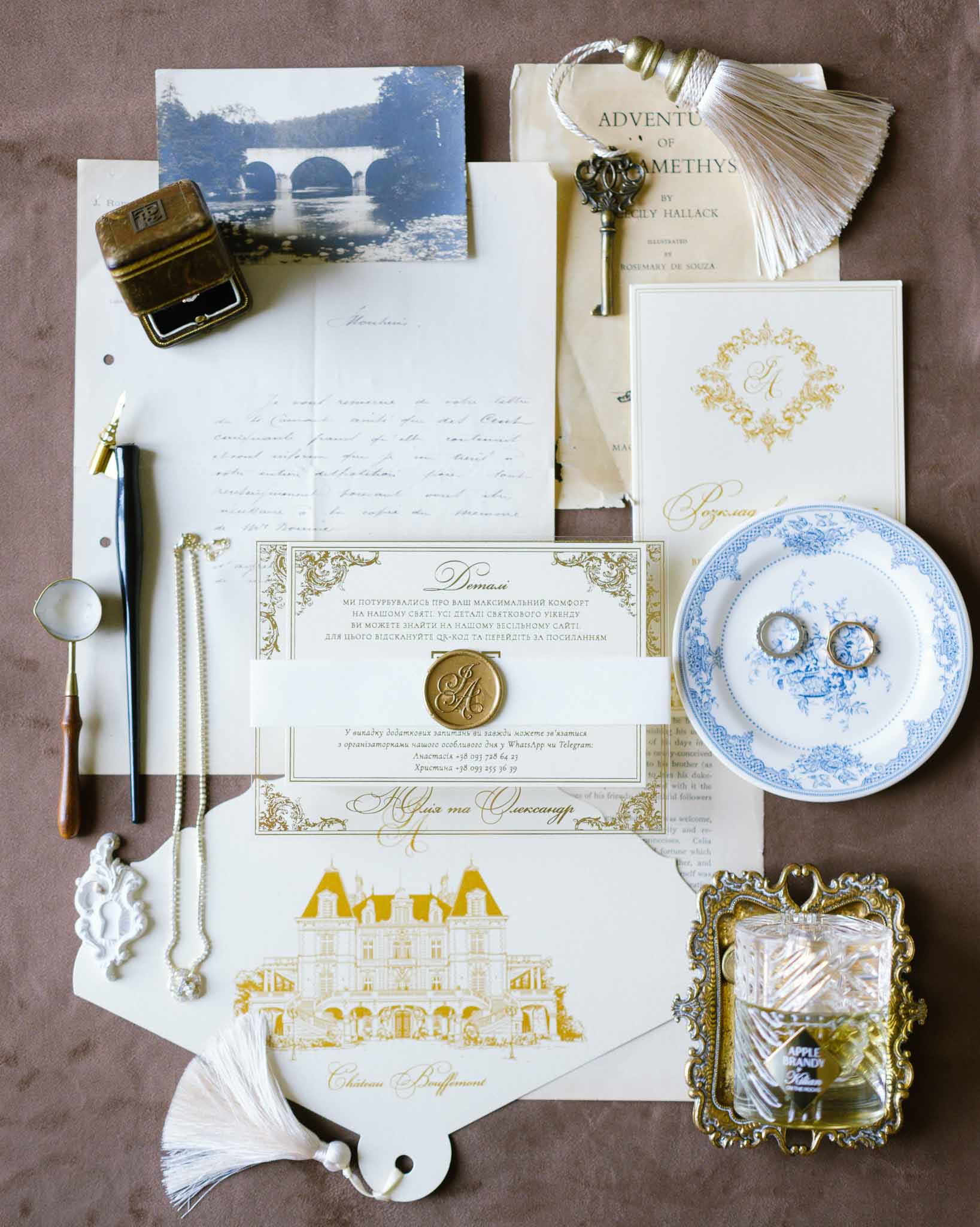Wedding stationery flat lay with gold foil chateau illustration, wax seal, rings, perfume, and vintage book