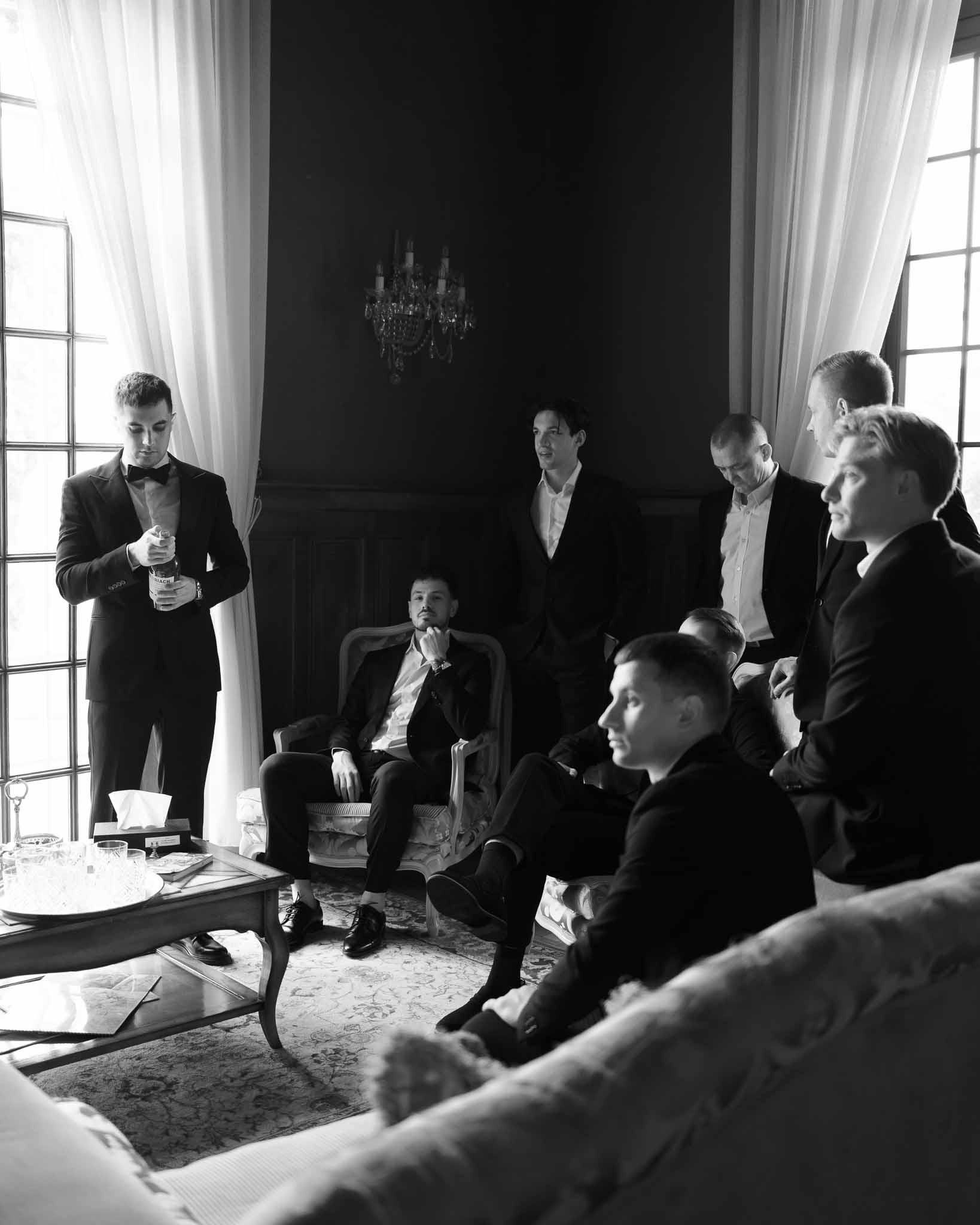 Black and white getting ready scene of groom and seven groomsmen in chateau salon with chandelier