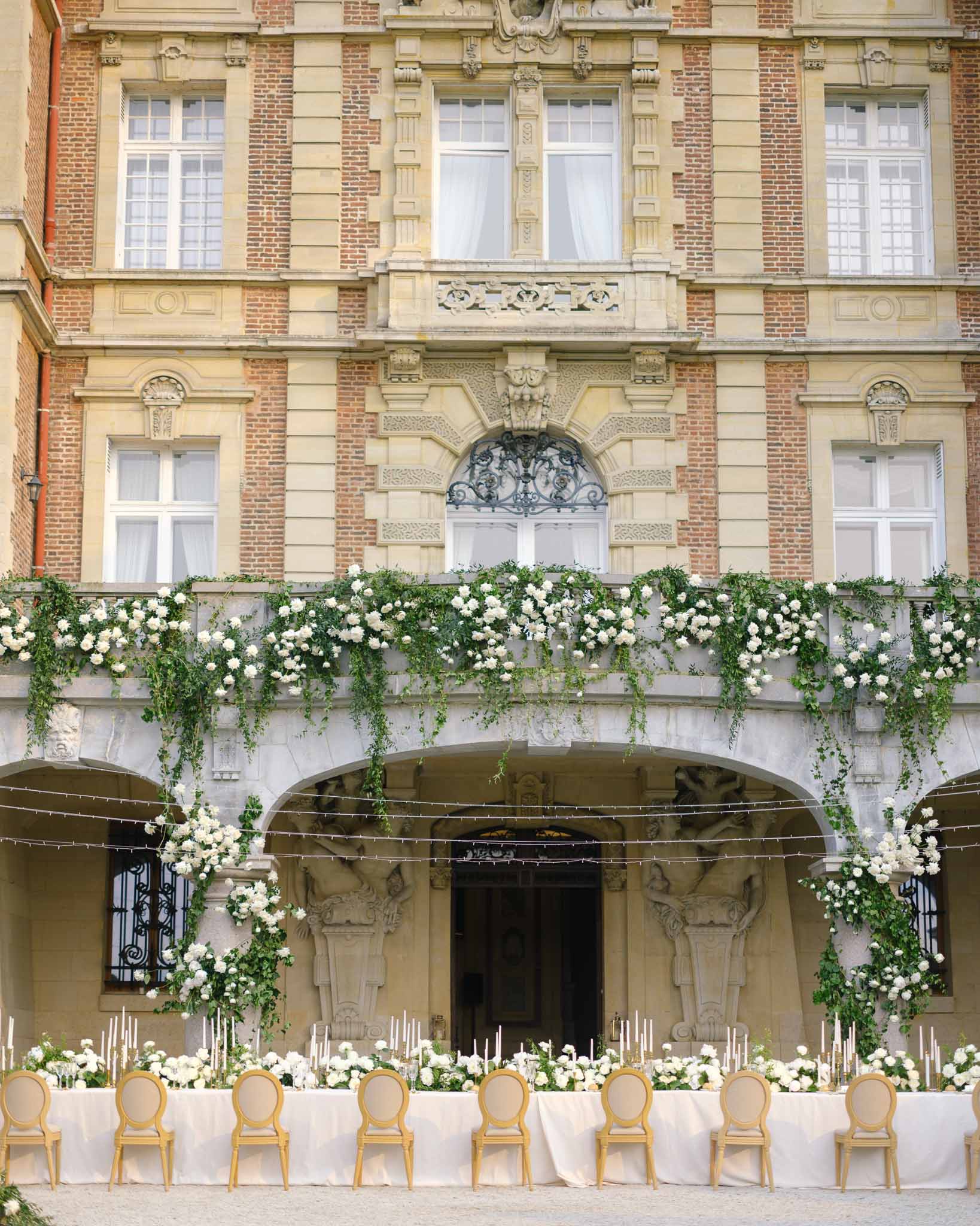 Long reception table with white roses and taper candles set in chateau courtyard with floral garland on balustrade