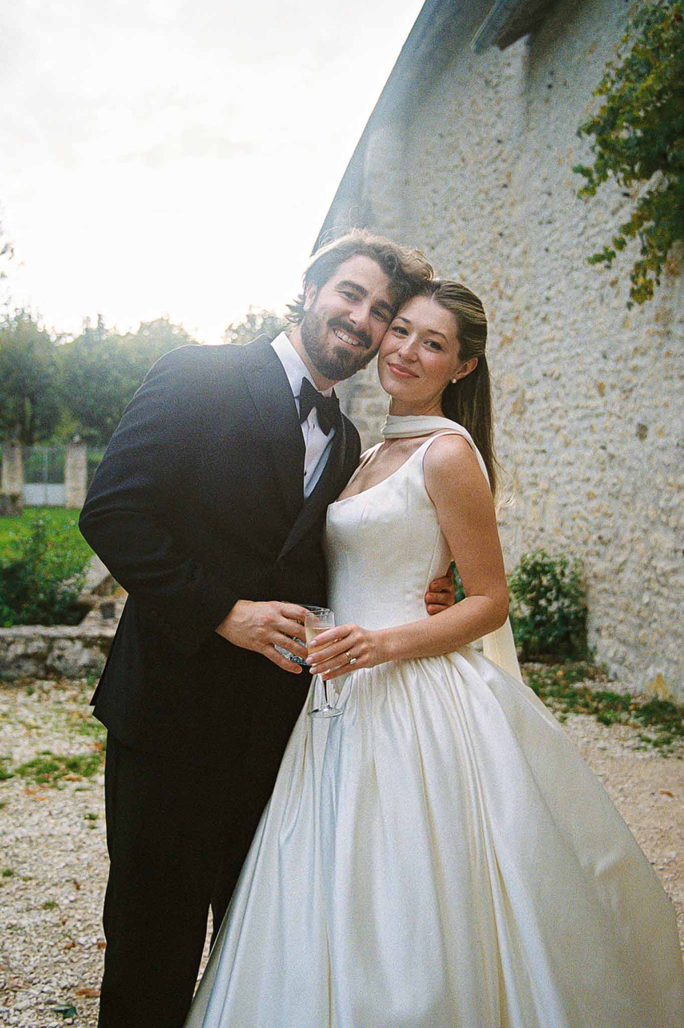 Bride in ivory satin ballgown and groom in black tuxedo sharing champagne on a chateau gravel courtyard in warm film tones