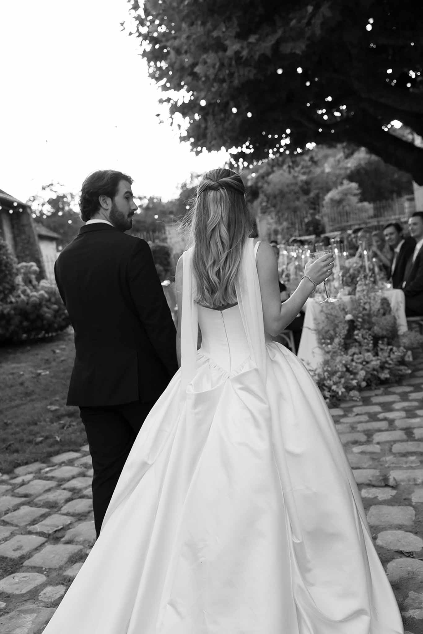 Black and white of couple from behind walking to reception table with fairy light canopy and champagne flute in hand