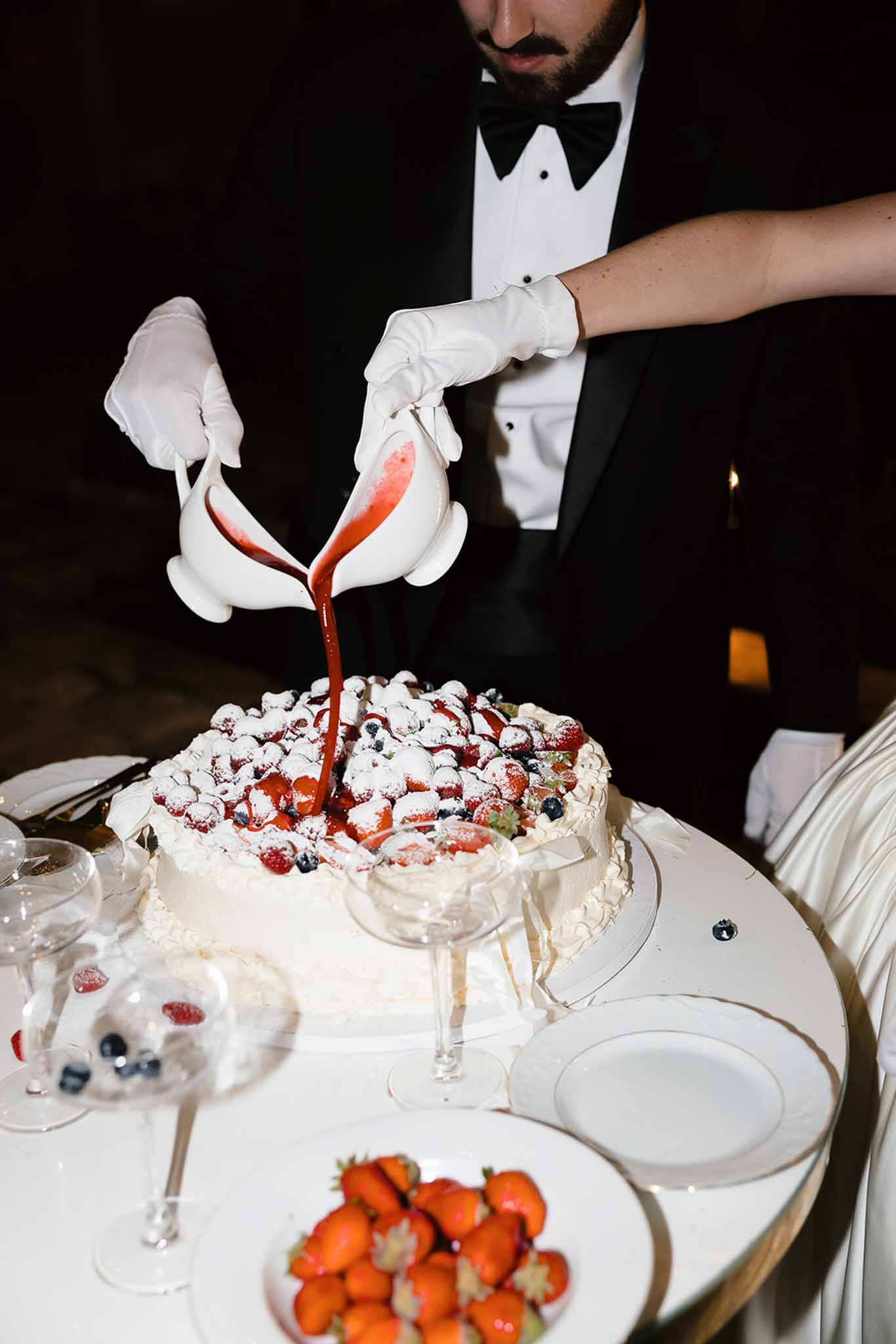 Waiter in tuxedo pouring berry coulis onto white wedding cake topped with strawberries and blueberries