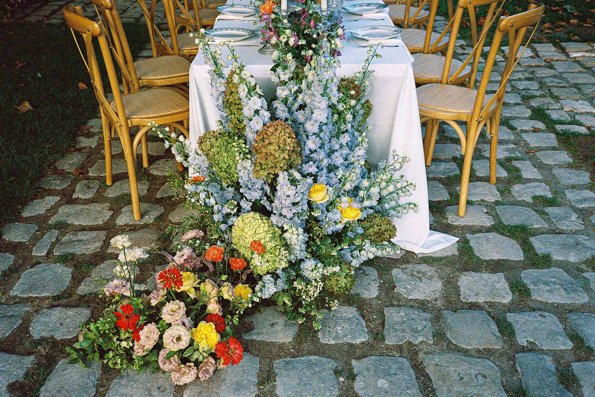Outdoor reception table with cascading floral centerpiece of delphiniums, roses, and zinnias on cobblestone