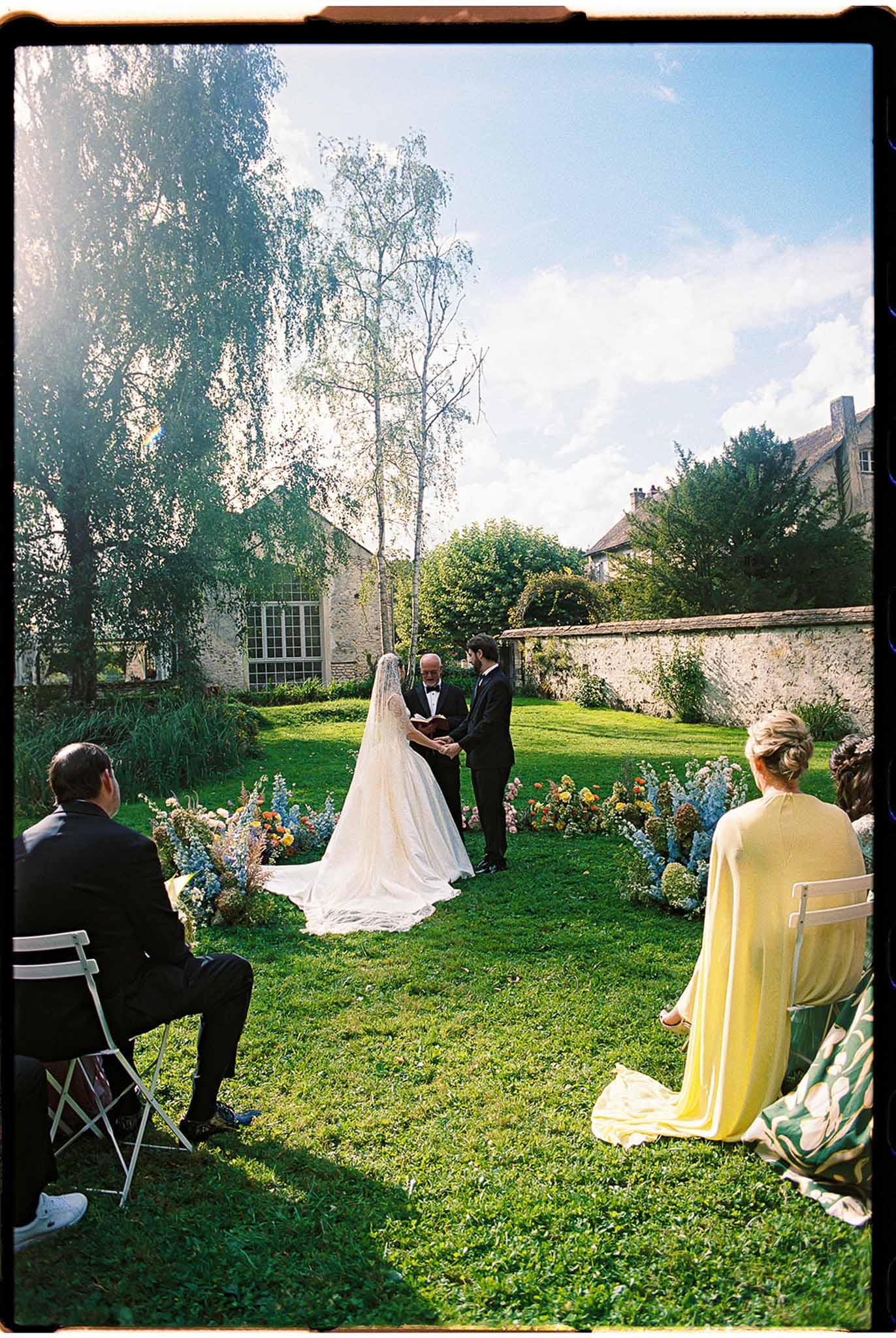 Outdoor ceremony on walled garden lawn with bride and groom at altar, wildflower aisle arrangements in mixed colours