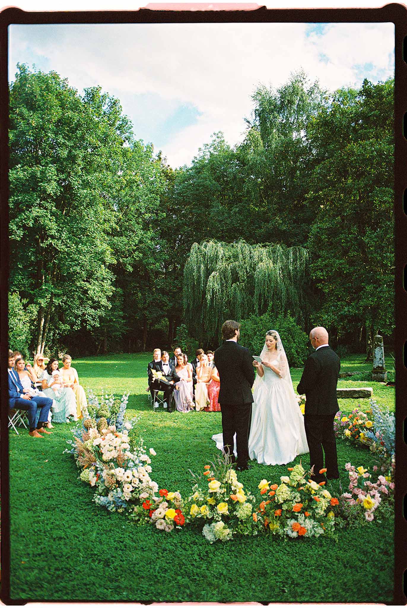 Outdoor ceremony on lawn with bride reading vows, ground-level floral arrangement of poppies and ranunculus