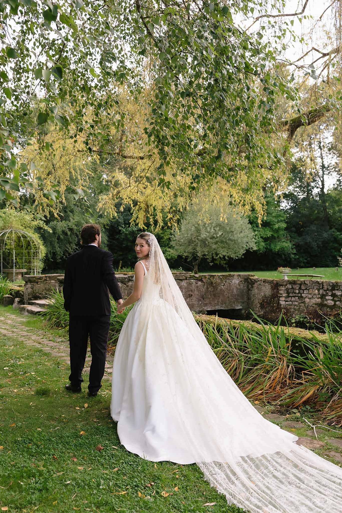 Couple walking through estate grounds, bride trailing cathedral lace veil across lawn under weeping birch
