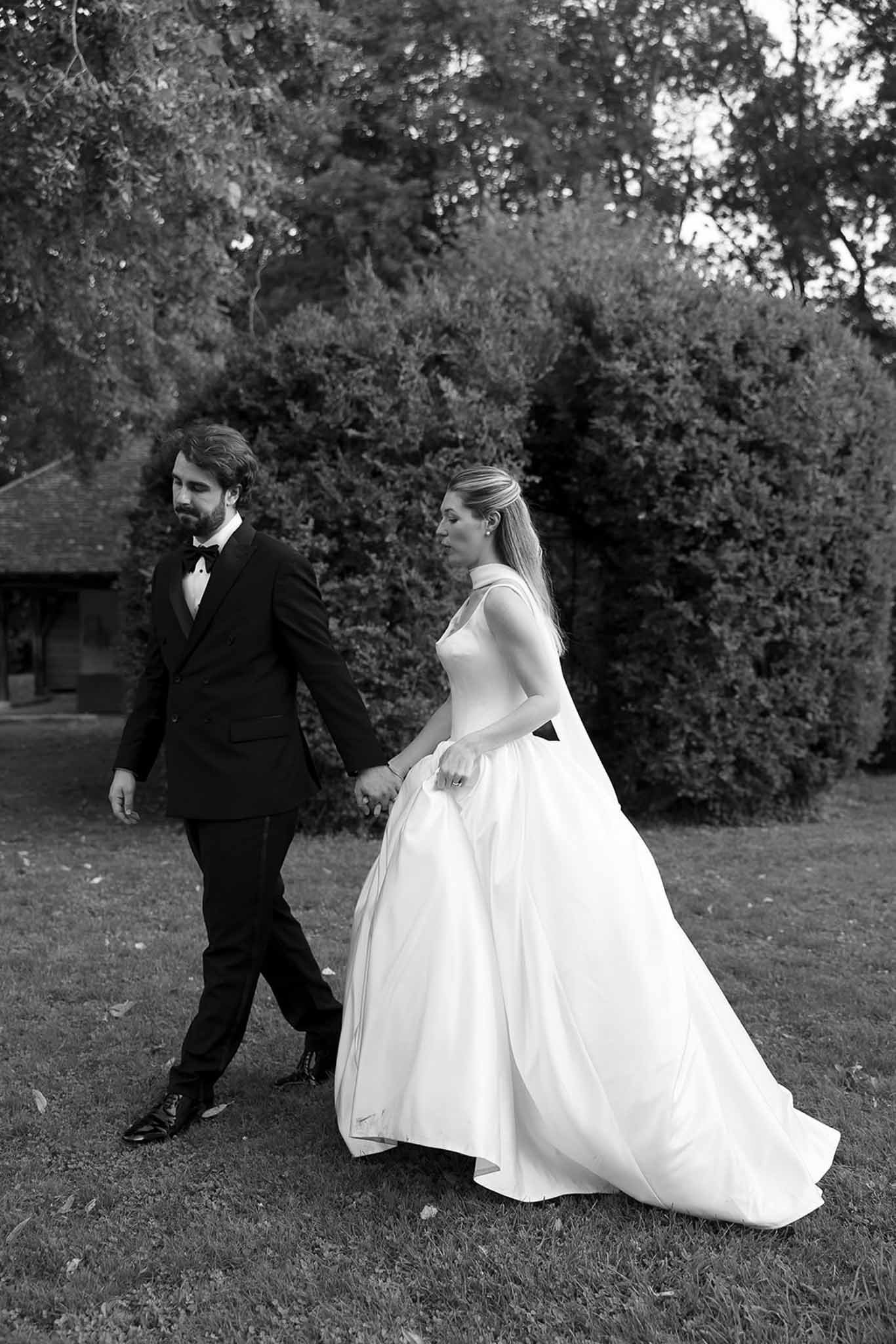 Black and white portrait of bride in halter ball gown and groom in tuxedo walking hand in hand across a lawn