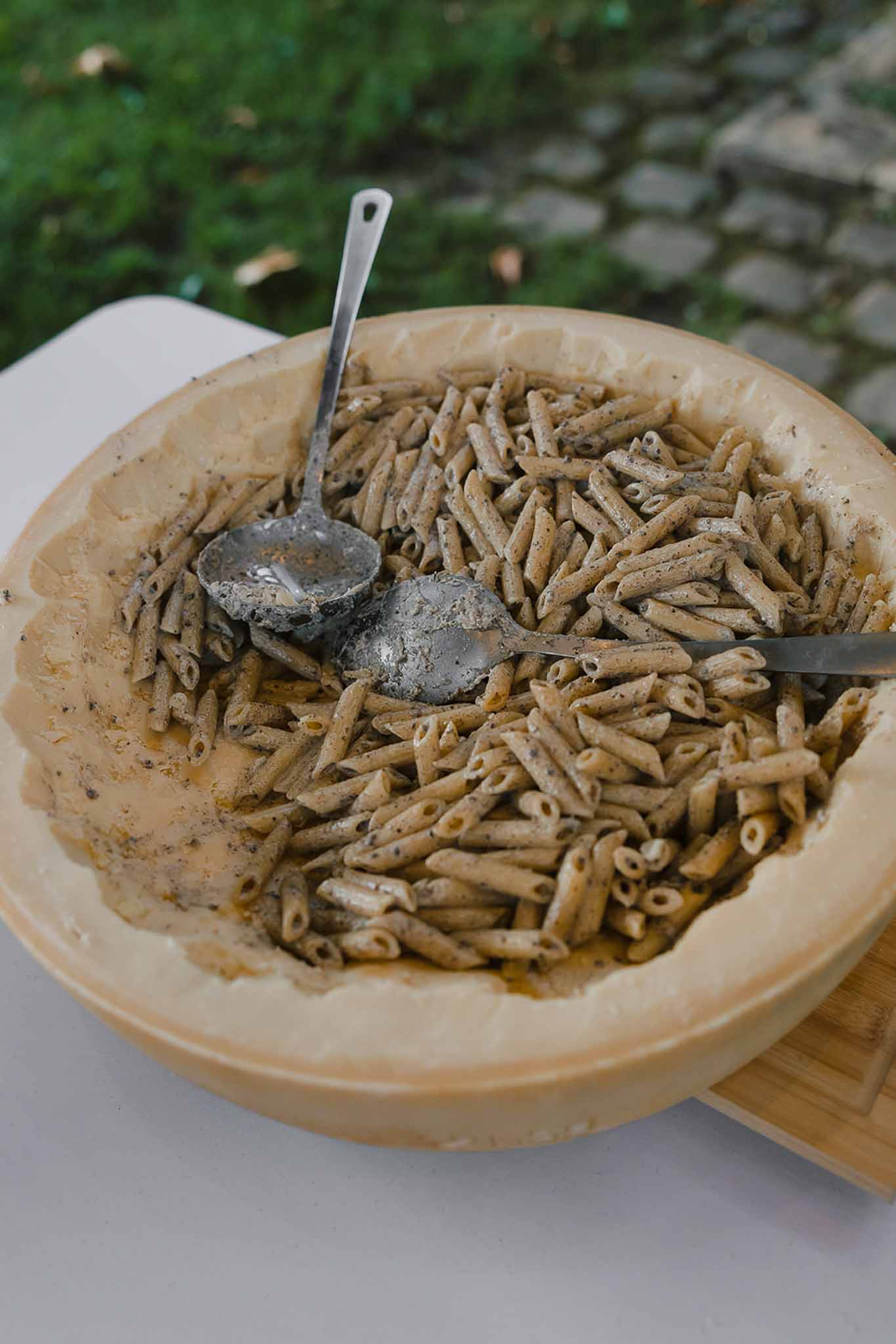 Penne pasta served from hollowed parmesan wheel with silver spoons at outdoor reception food station