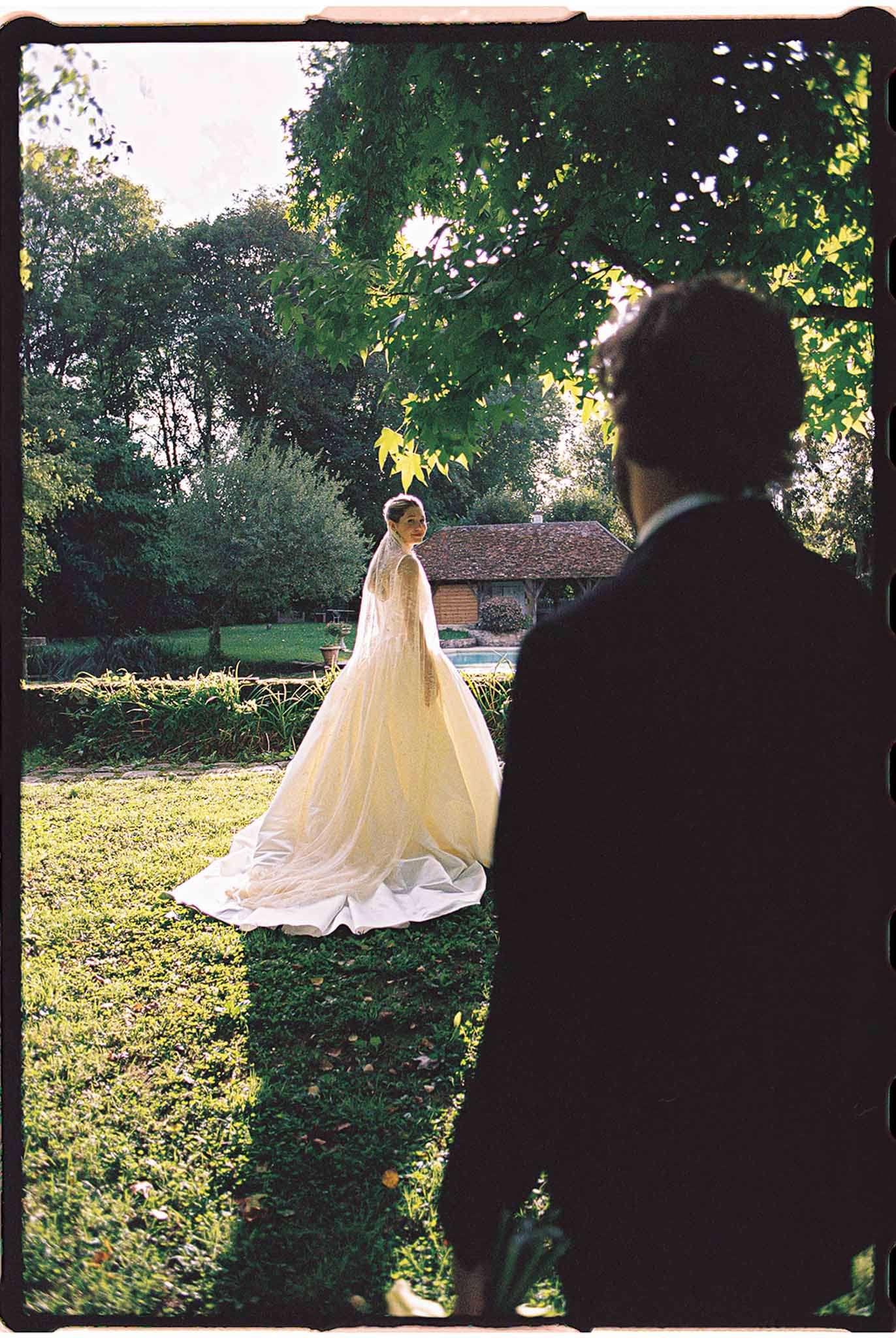 Bride in ivory ball gown with cathedral-length veil on lawn, groom in dark suit seen from behind at country estate