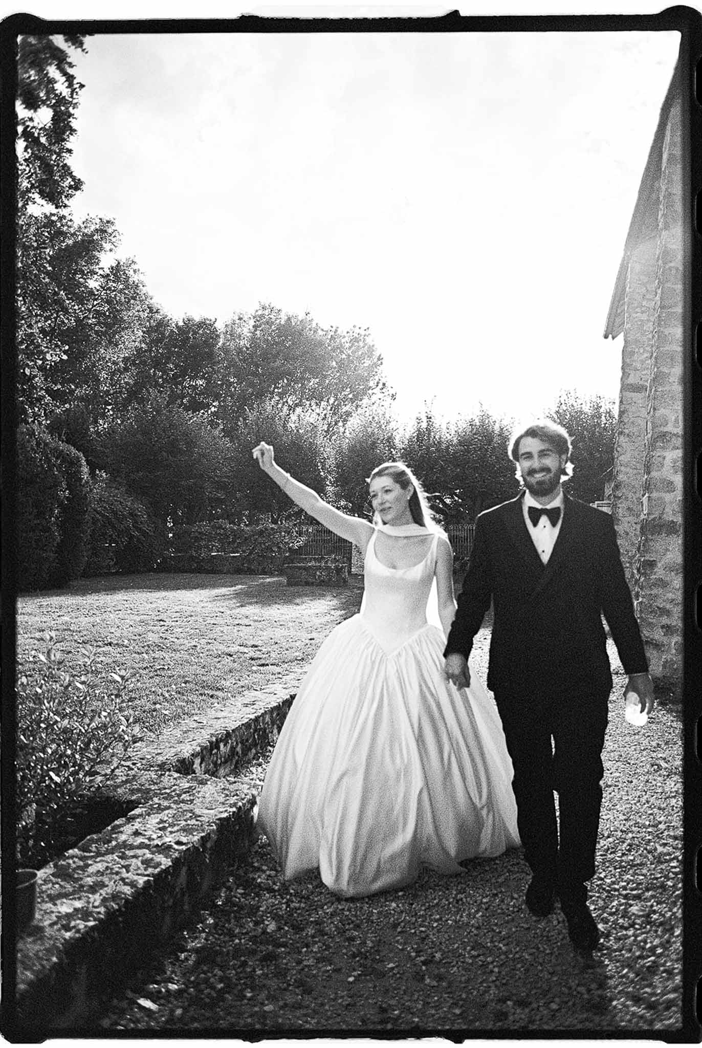 Black-and-white film photo of bride in ball gown raising her arm and groom in suit walking along a gravel path