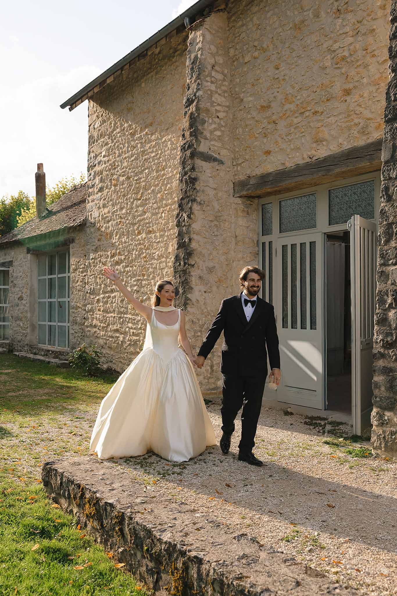 Bride waving in ivory ballgown walking with groom in tuxedo along stone building in warm golden afternoon light