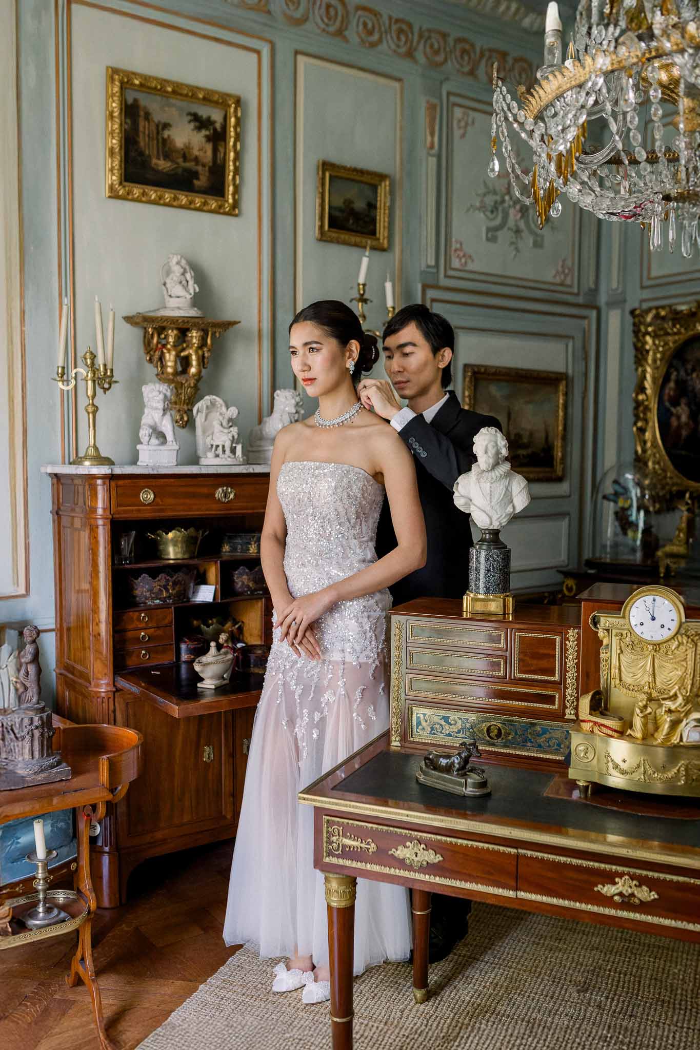 Groom adjusting bride's necklace in chateau salon with blue-grey panels gilt paintings and crystal chandelier